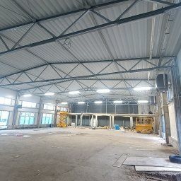 KS DACH SYSTEM DARIA ŚLUBOWSKA - Wnętrze hali przemysłowej z widoczną konstrukcją dachu z blachy trapezowej i stalowymi dźwigarami. Na podłodze widoczne ślady prac budowlanych i podnośniki koszowe.