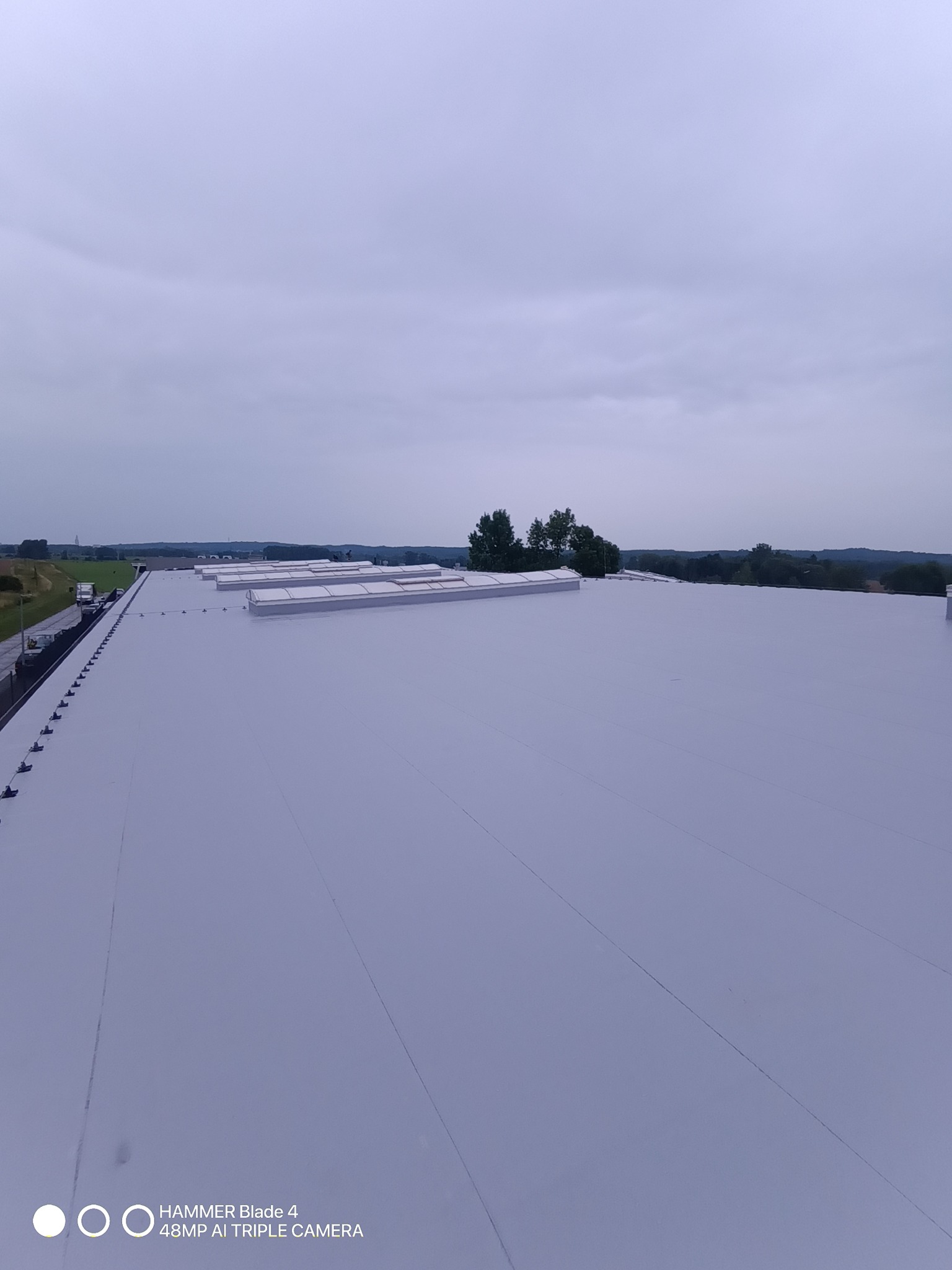 Jasnoszary, płaski dach przemysłowy z świetlikami, widziany z góry, z zielonym krajobrazem w tle pod pochmurnym niebem. Widoczne zabezpieczenia przeciwśniegowe.