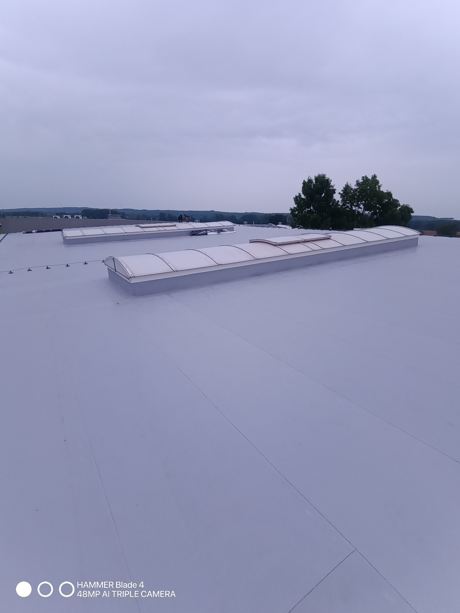 Jasnoszary dach płaski z dwoma świetlikami kopułowymi. W tle zieleń drzew i zabudowania. Widok z góry, pochmurne niebo.