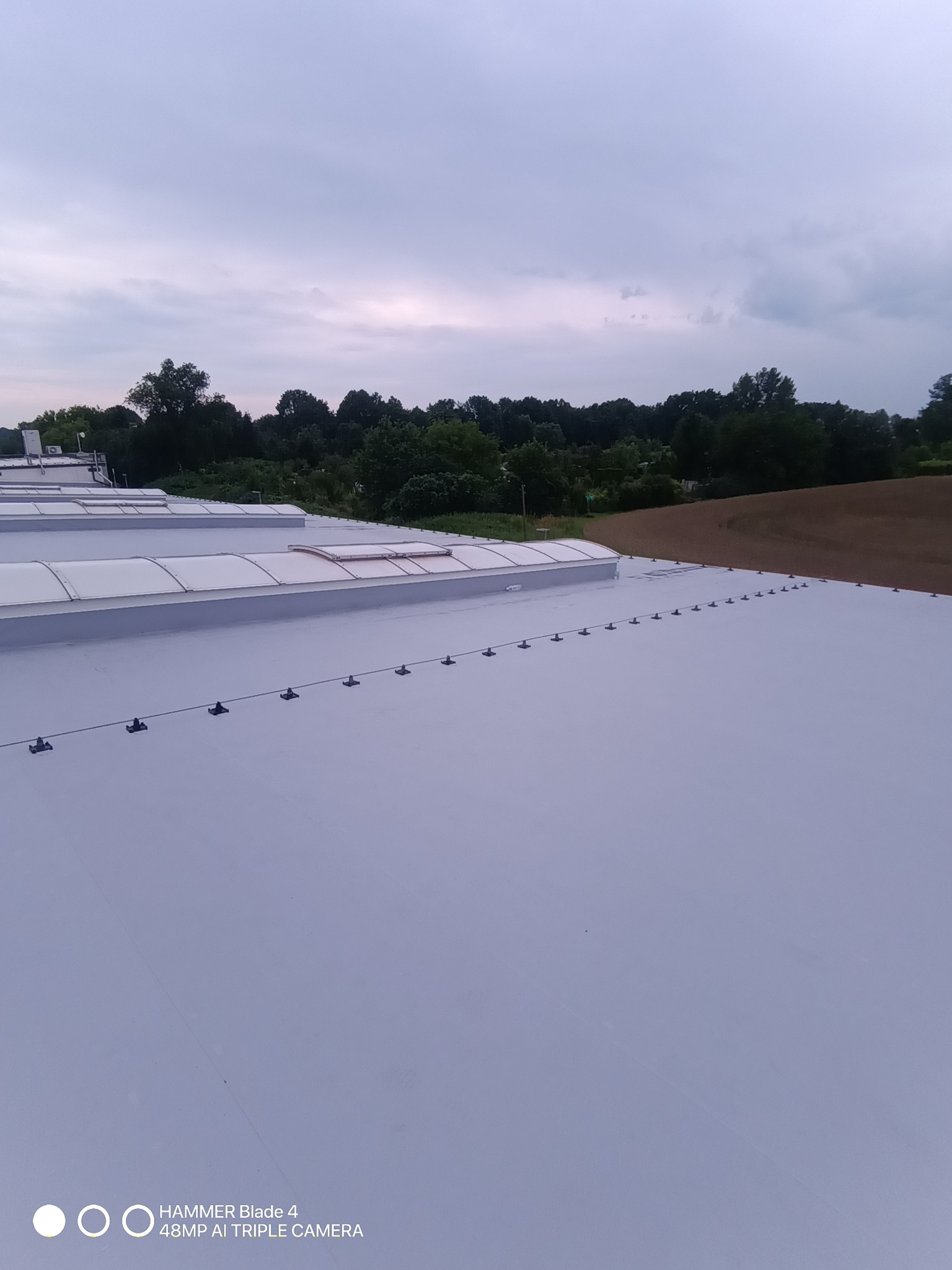 Jasnoszary dach przemysłowy z świetlikami i systemem odgromowym na tle zieleni i pochmurnego nieba. Widok z góry, szeroki kadr.