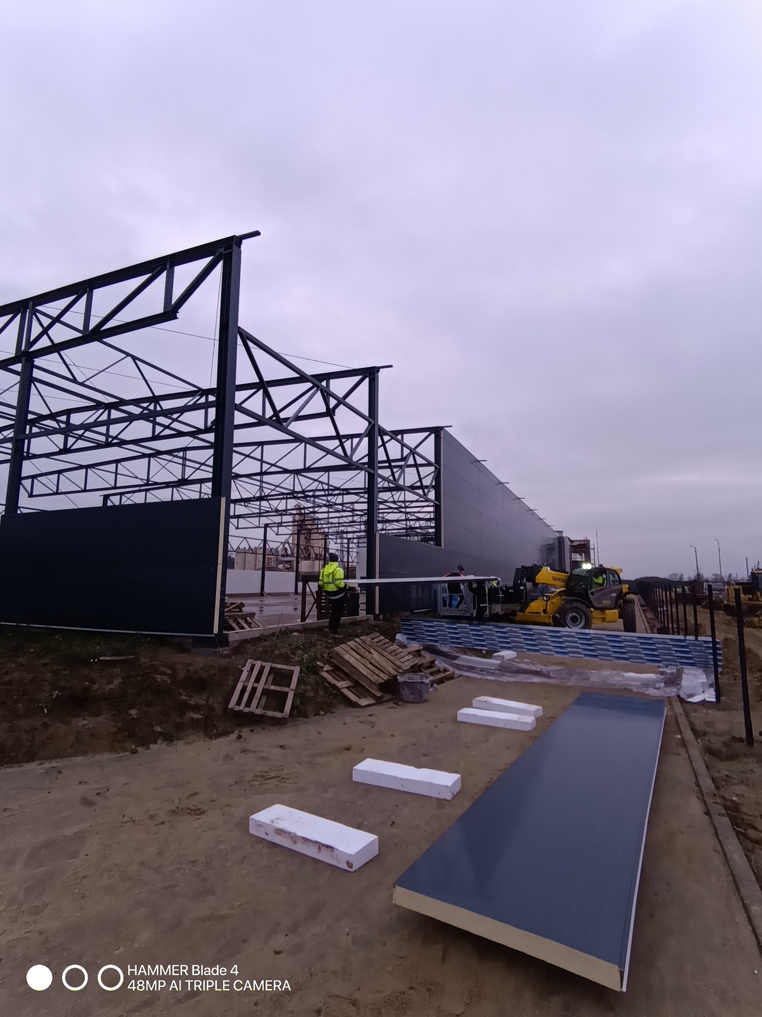 Montaż hali z paneli warstwowych. Widoczny stalowy szkielet konstrukcji, pracownicy i podnośnik teleskopowy. Na pierwszym planie leżą panele.