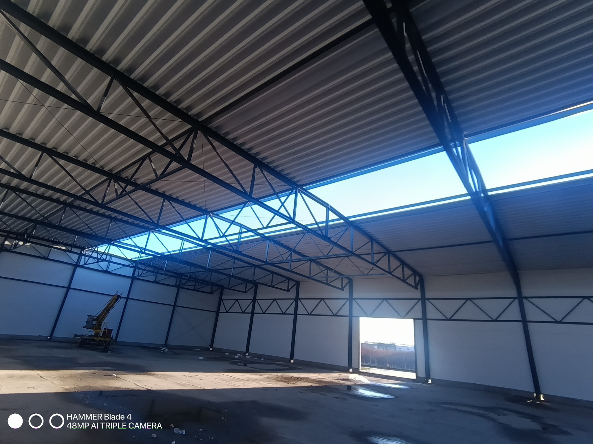 Wnętrze hali z widoczną konstrukcją dachu z blachy trapezowej i stalowymi elementami nośnymi. W tle widoczny sprzęt budowlany i wjazd do hali.