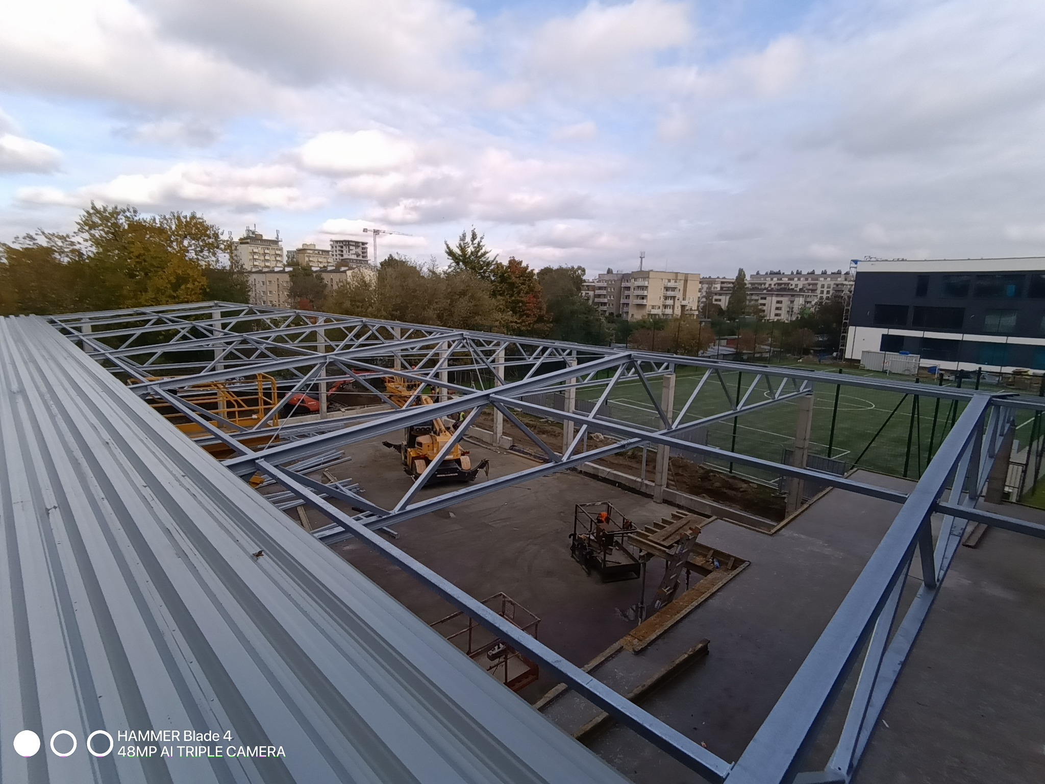 Konstrukcja stalowa dachu w trakcie montażu, widok z góry na plac budowy z maszynami i elementami konstrukcyjnymi na tle zabudowy miejskiej i zieleni.
