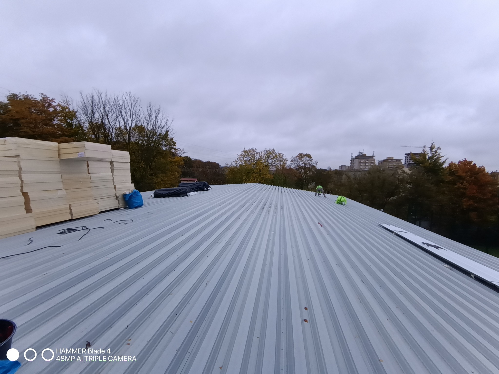 Montaż blachy trapezowej na dachu z widocznym ociepleniem. Prace dekarskie w toku, widok z góry na szary dach i pracowników w oddali.