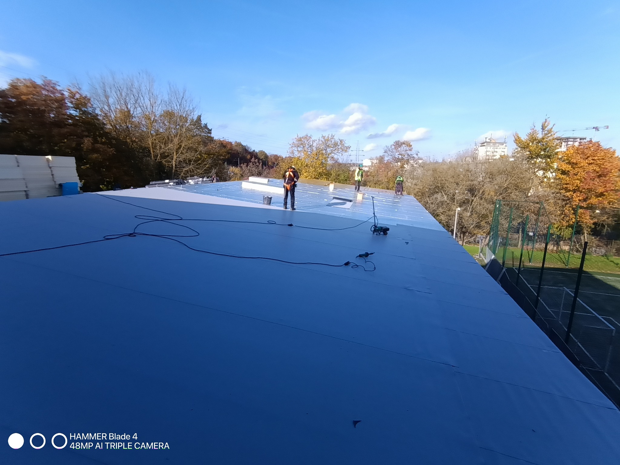 Ekipa w pracy na płaskim dachu pokrytym szarą membraną, widoczne narzędzia i materiały izolacyjne. W tle drzewa i budynki w oddali.