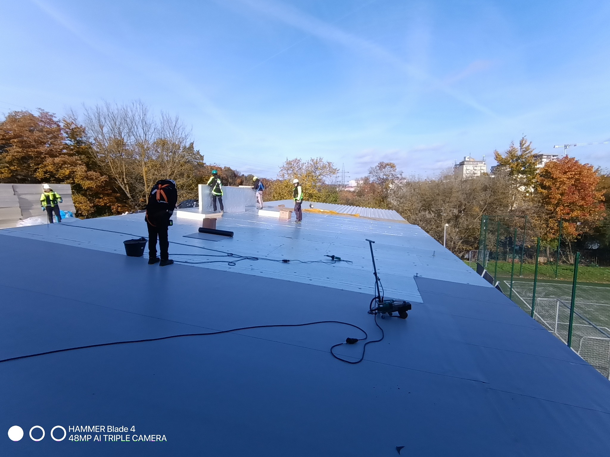 Prace na dachu: ekipa w kaskach montuje izolację termiczną na płaskim dachu budynku, widoczne narzędzia i materiały izolacyjne. W tle drzewa i budynki.