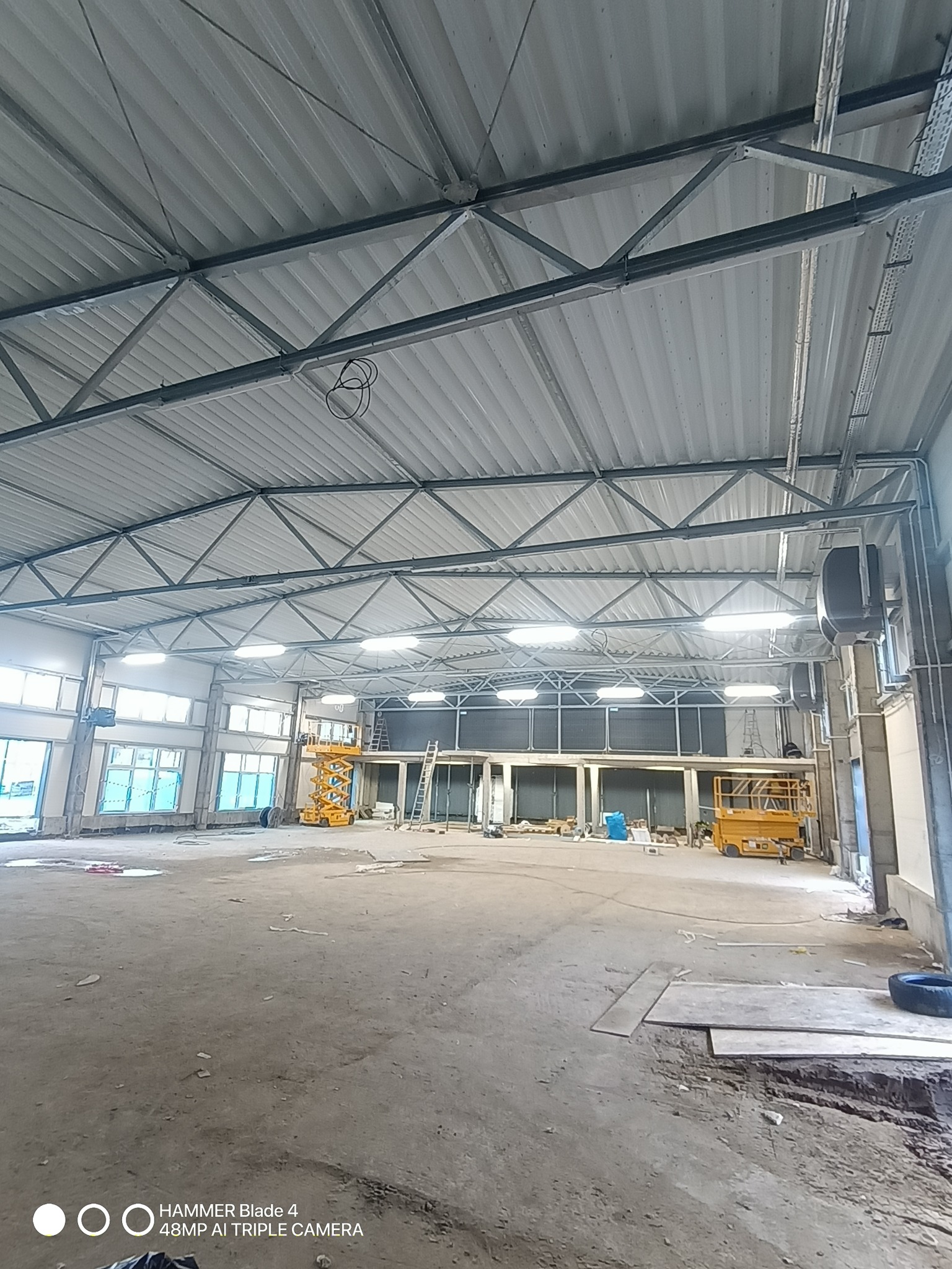 Wnętrze hali przemysłowej z widoczną konstrukcją dachu z blachy trapezowej i stalowymi dźwigarami. Na podłodze widoczne ślady prac budowlanych i podnośniki koszowe.