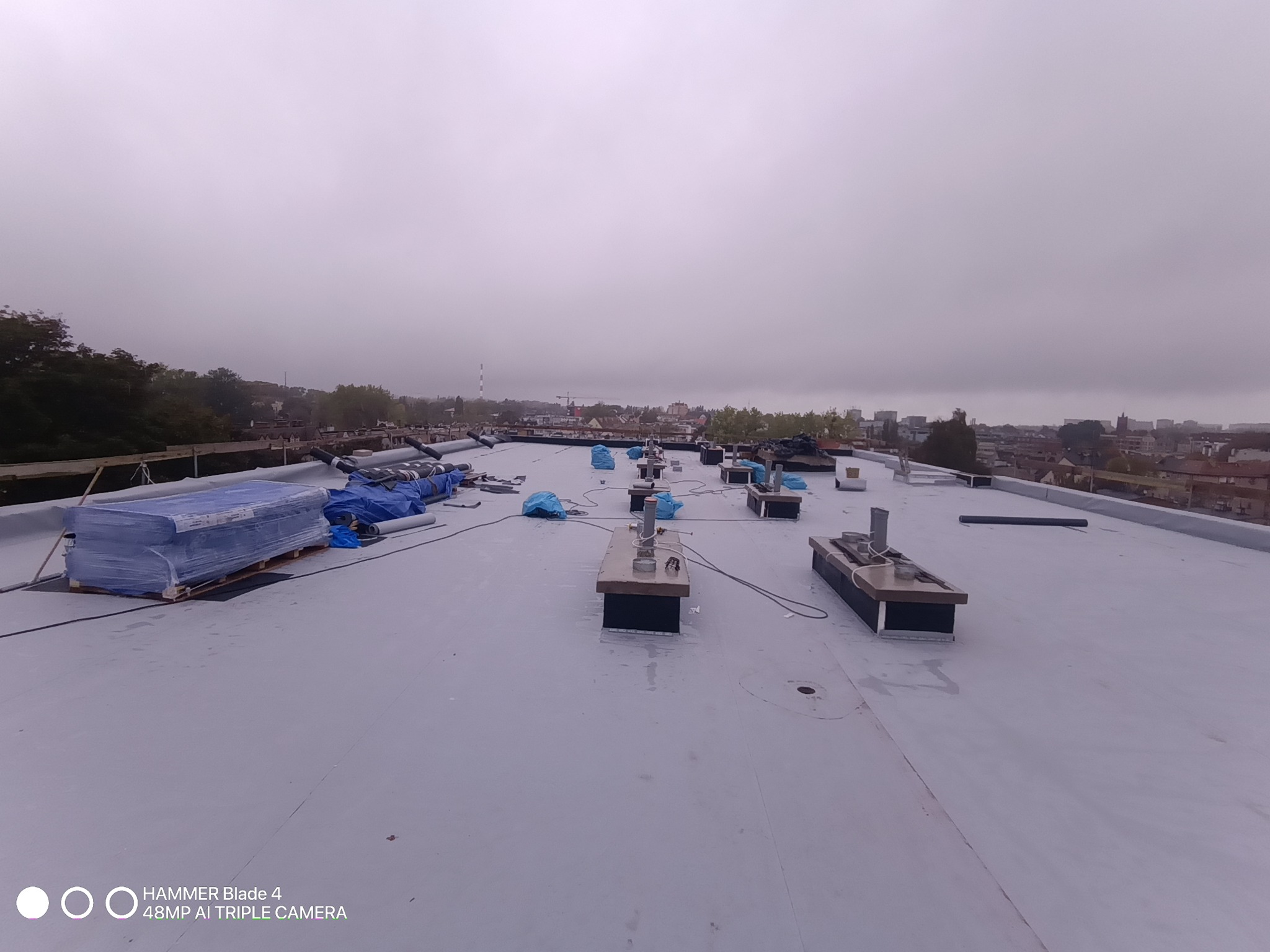 Szary dach płaski z elementami wentylacyjnymi i zapasem materiałów budowlanych w tle zabudowa miejska w pochmurny dzień.