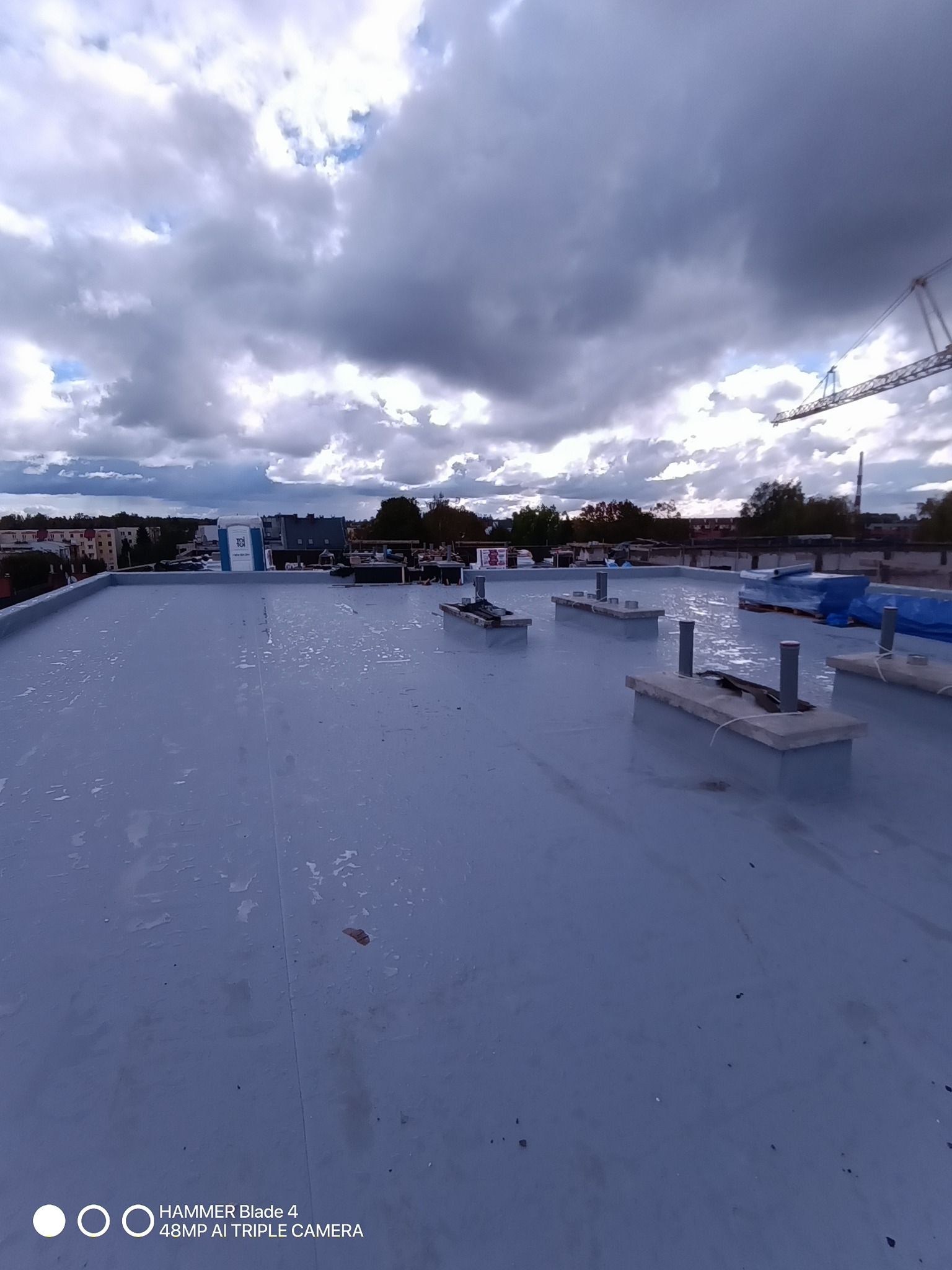 Szary, płaski dach budynku z wystającymi elementami konstrukcyjnymi i widokiem na pochmurne niebo z dźwigiem w tle. Widoczne ślady po deszczu.