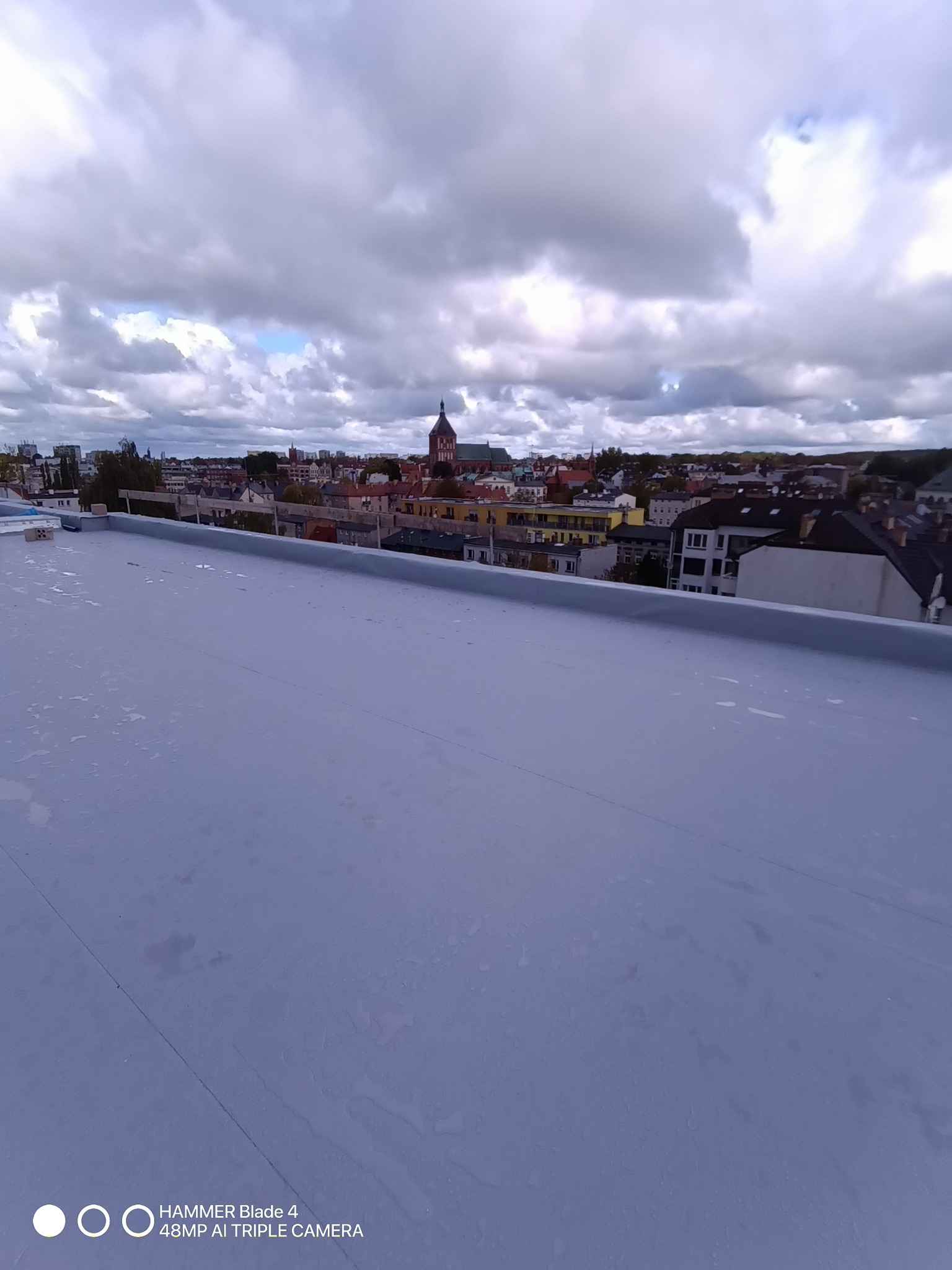 Nowe, szare pokrycie dachu płaskiego z widokiem na miasto i kościół w tle, wykonane w pochmurny dzień. Widoczne ślady po deszczu na powierzchni.