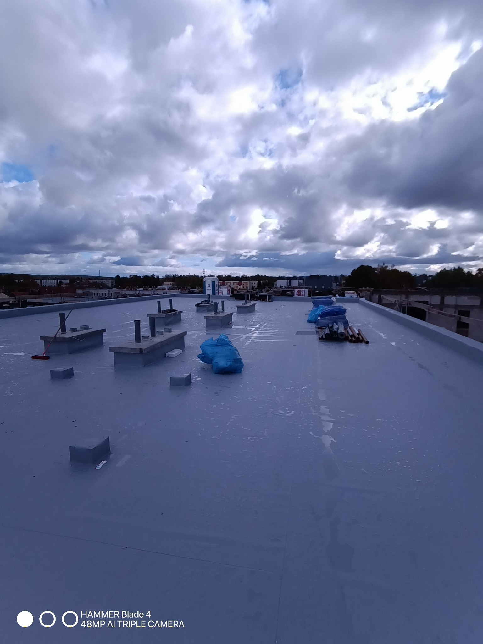 Szary, płaski dach po hydroizolacji z wystającymi elementami konstrukcyjnymi i niebieskimi workami. W tle zabudowa miejska i pochmurne niebo.