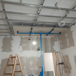 Sylwester Bieniecki - Montaż sufitu podwieszanego z płyt karton-gips. Widoczne profile, płyta na podnośniku, szpachlowane ściany i drabiny. Prace wykończeniowe w trakcie realizacji.