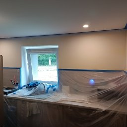 Sylwester Bieniecki - Kuchnia w trakcie malowania: ściany zabezpieczone folią i taśmą malarską, widoczne narzędzia malarskie, sufit z oświetleniem punktowym, przygotowania do malowania.