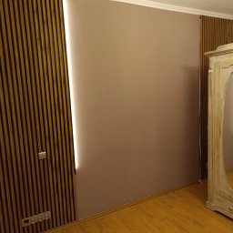 Sylwester Bieniecki - Beżowa ściana z drewnianymi lamelami i ukrytym oświetleniem LED. Widoczny fragment podłogi i szafy. Nowoczesny design wnętrza.