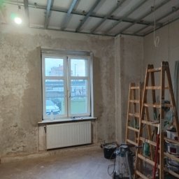 Sylwester Bieniecki - Wnętrze w trakcie prac w Kłodzku: surowe ściany, metalowa konstrukcja sufitu podwieszanego, drabiny, odkurzacz przemysłowy. Widok na okno i budynek na zewnątrz.