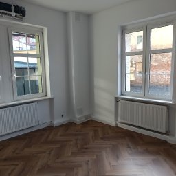 Sylwester Bieniecki - Jasne wnętrze po remoncie: parkiet w jodełkę, białe ściany, okna z grzejnikami. Widoczne niedokończone detale instalacji elektrycznej i akcesoria montażowe.