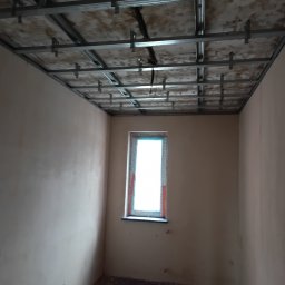 Sylwester Bieniecki - Pokój w trakcie montażu sufitu podwieszanego z metalowej konstrukcji. Widoczne profile i elementy mocujące. Ściany otynkowane, okno z widokiem na zewnątrz.