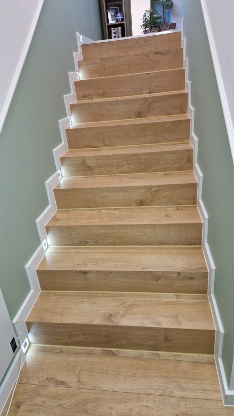 Drewniane schody z subtelnym oświetleniem LED wzdłuż krawędzi, prowadzące w górę, na tle ściany w odcieniach zieleni i bieli. Widok z dołu.