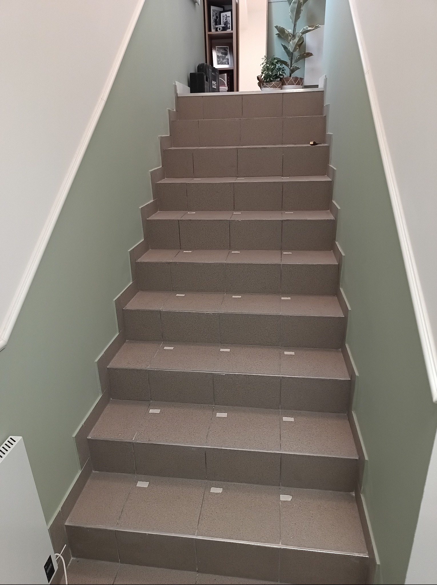 Wykończone schody wewnątrz budynku, wyłożone brązowymi płytkami z delikatnym wzorem. Ściany pomalowane na jasny, pastelowy odcień zieleni. Widoczny grzejnik i fragment regału.