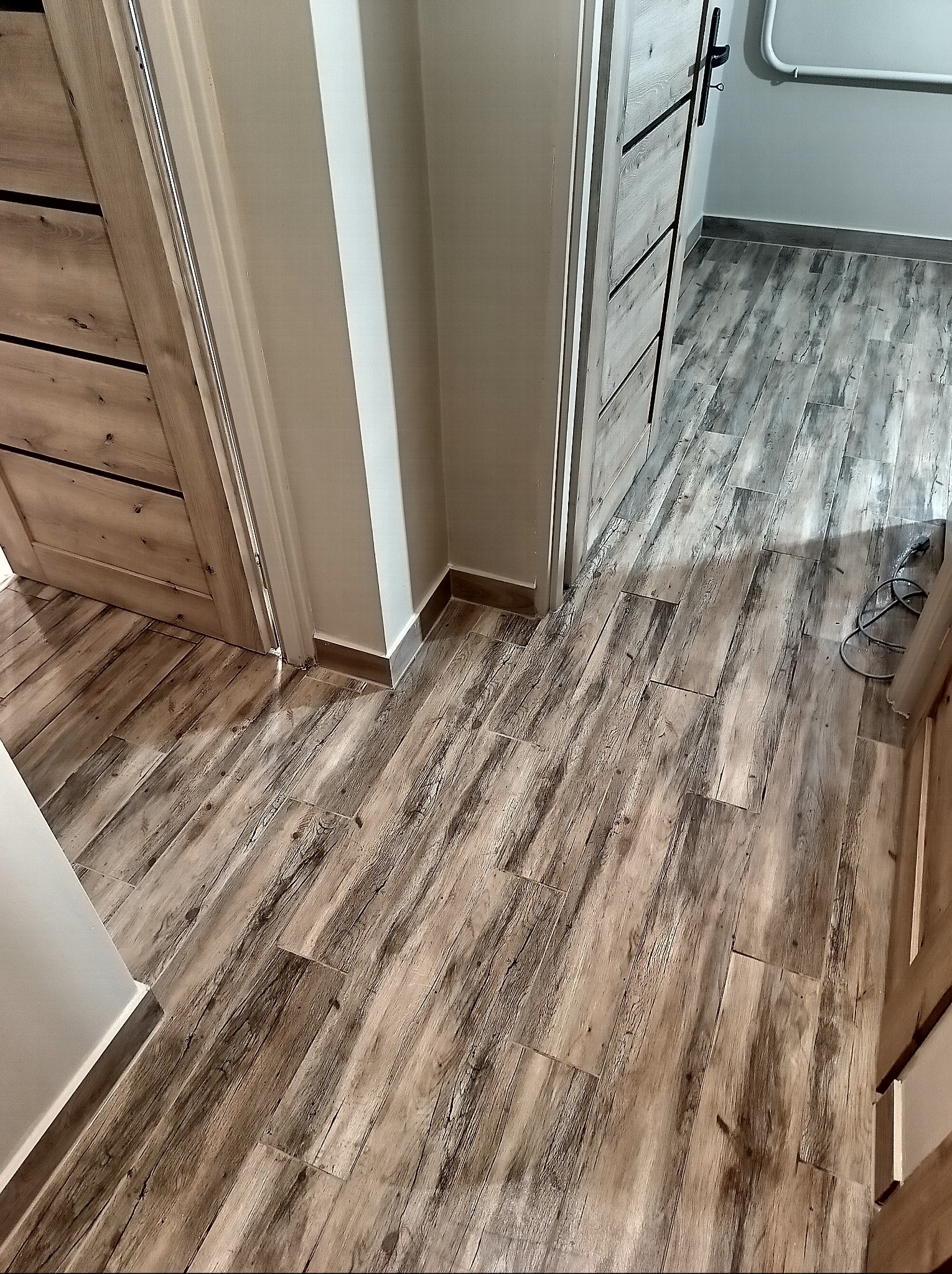 Ułożona podłoga z płytek imitujących drewno w korytarzu, widoczne dwie pary drzwi z ościeżnicami. Płytki ułożone wzdłuż korytarza, z widocznymi fugami.