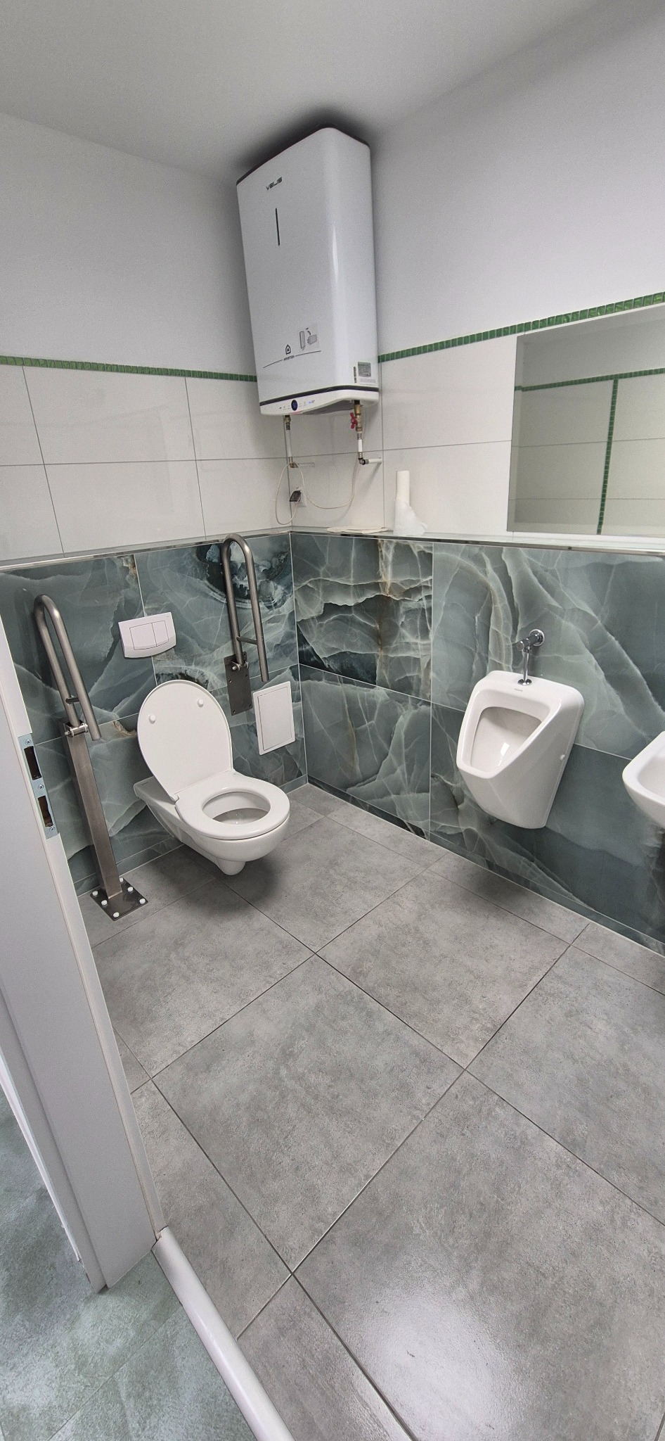 Wykończona łazienka publiczna z toaletą dla niepełnosprawnych, pisuarami, szarą podłogą i płytkami imitującymi marmur na ścianach. Widoczny bojler i uchwyty.