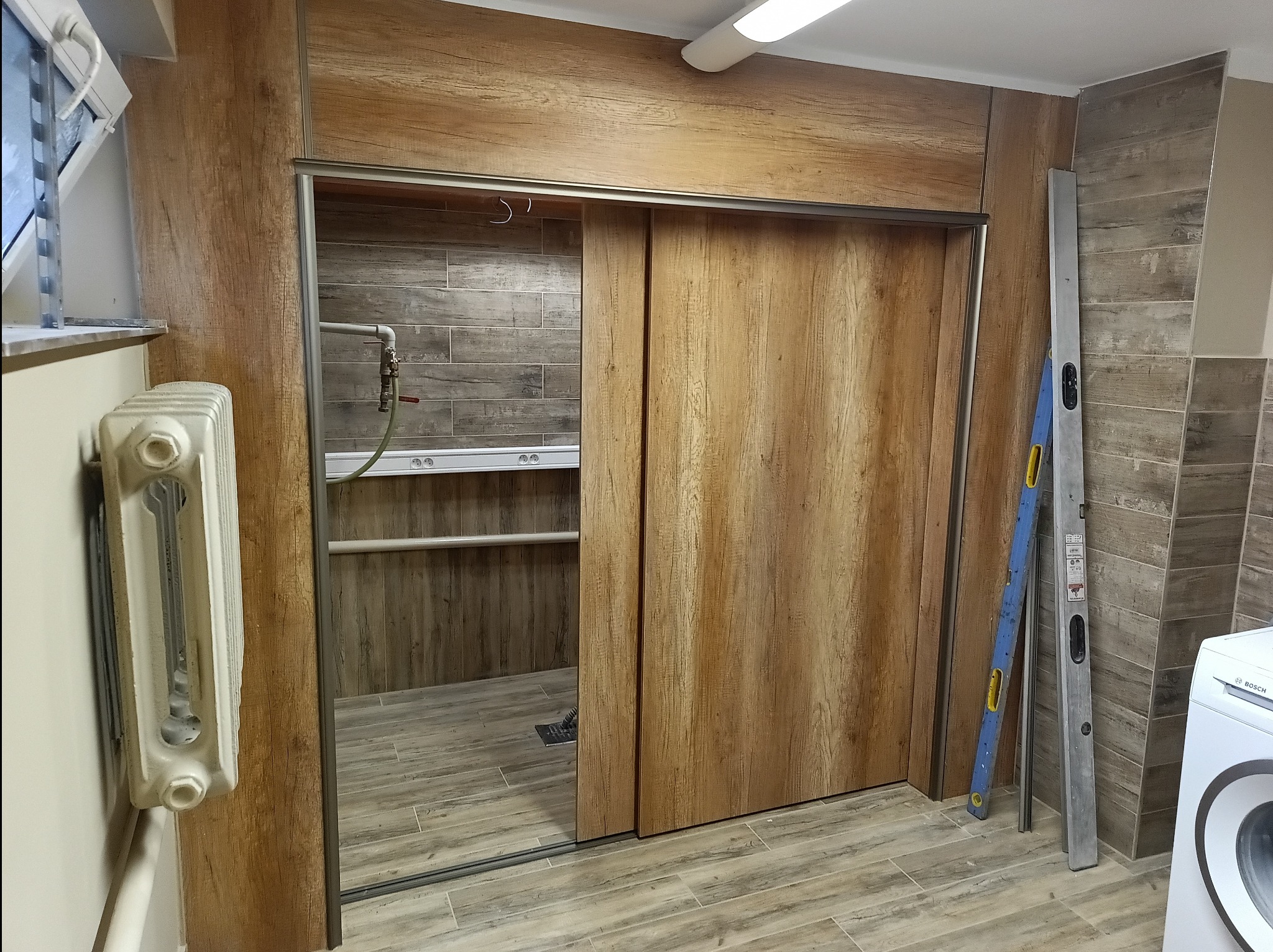 Wykończona łazienka w Kłodzku z przesuwnymi drzwiami o drewnianej fakturze, grzejnikiem i poziomnicami opartymi o ścianę z płytek imitujących drewno.