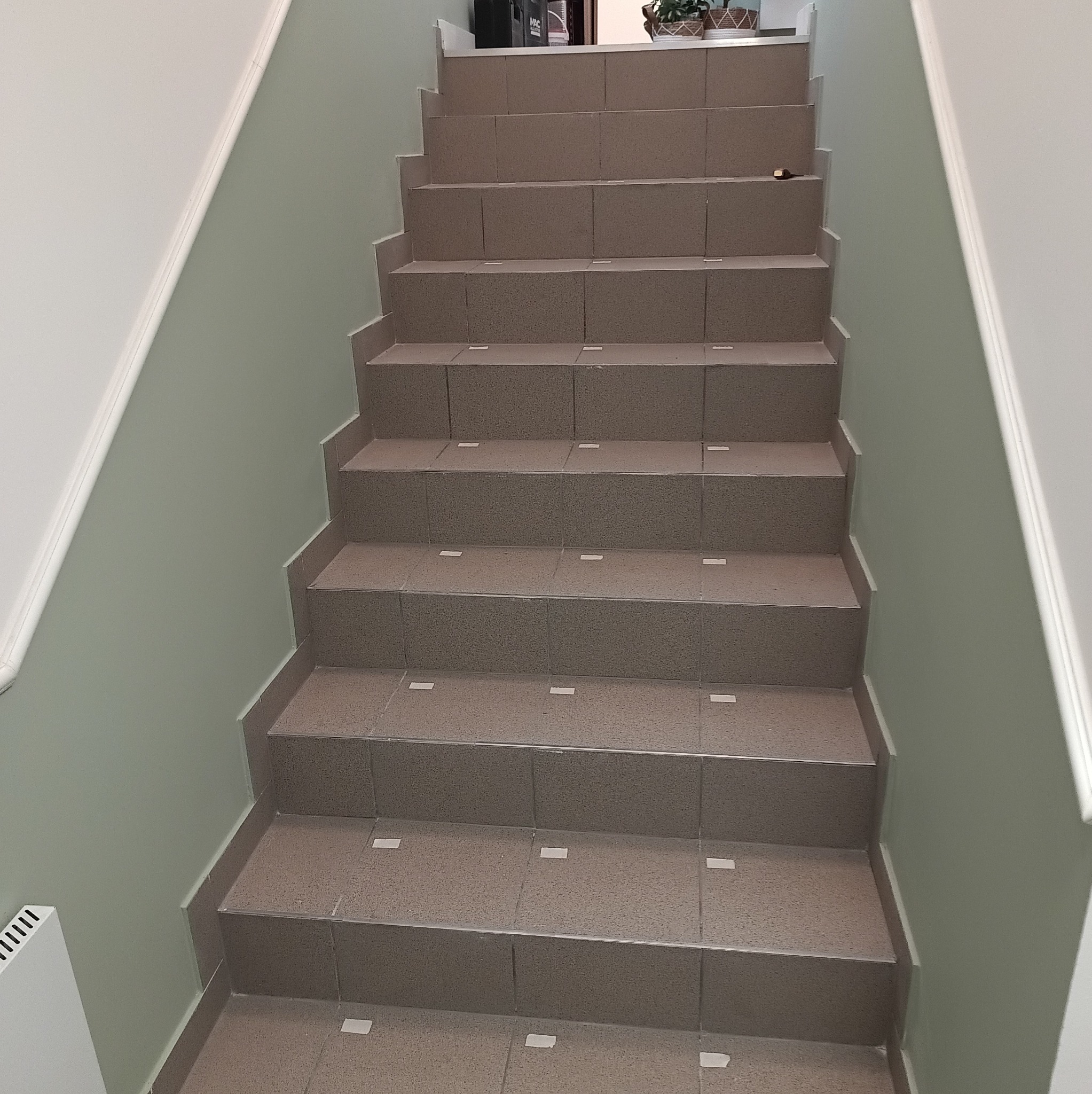 Wykończone schody z brązowych płytek, prowadzące w górę. Ściany pomalowane na jasnozielony kolor, z białymi listwami przypodłogowymi. Widoczny fragment grzejnika.