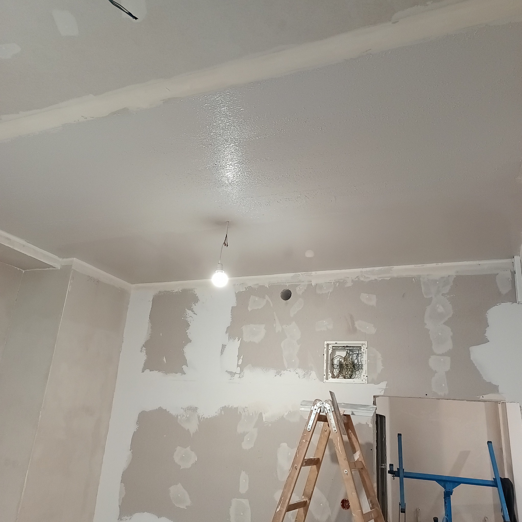 Pokój w trakcie prac wykończeniowych: częściowo wyszpachlowane ściany, sufit po pierwszym malowaniu, drabina i podnośnik do płyt. Widoczne kable i puszka elektryczna.