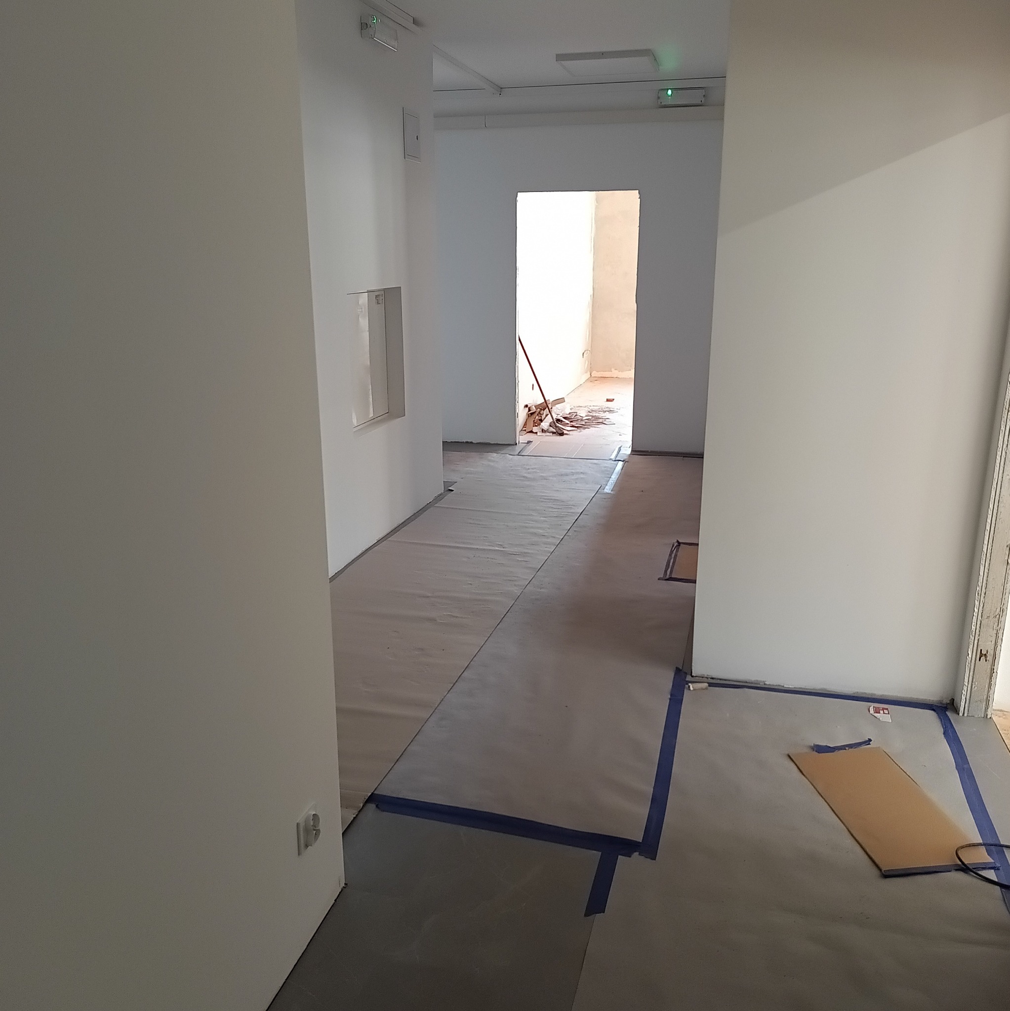 Białe ściany w korytarzu w trakcie prac wykończeniowych, podłoga zabezpieczona folią i taśmą malarską. Widoczne narzędzia i resztki materiałów budowlanych.