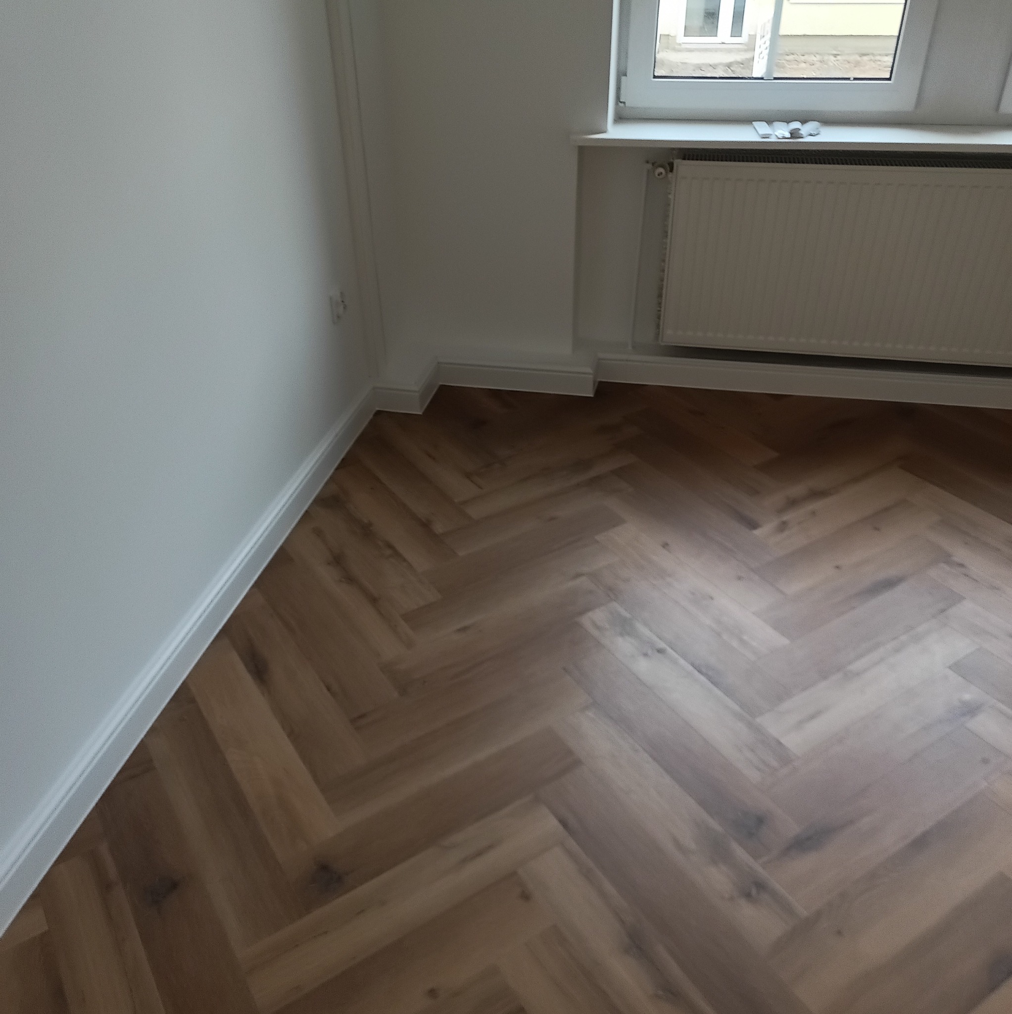 Parkiet ułożony w jodełkę w Kłodzku, widok na fragment pokoju z grzejnikiem i oknem. Jasne ściany i listwy przypodłogowe.