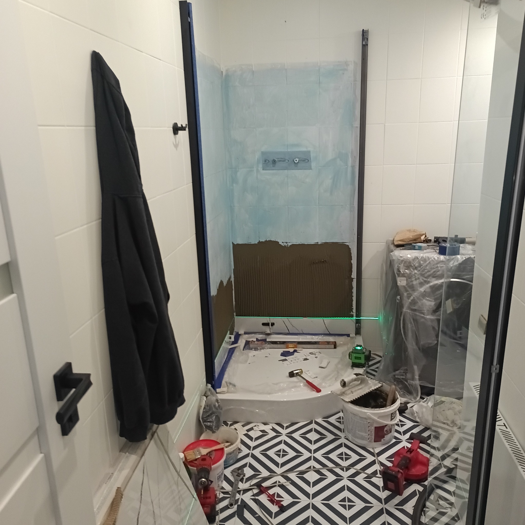 Remont łazienki: montaż płytek w kabinie prysznicowej z użyciem poziomicy laserowej. Widoczne narzędzia i materiały budowlane na podłodze w geometryczne wzory.