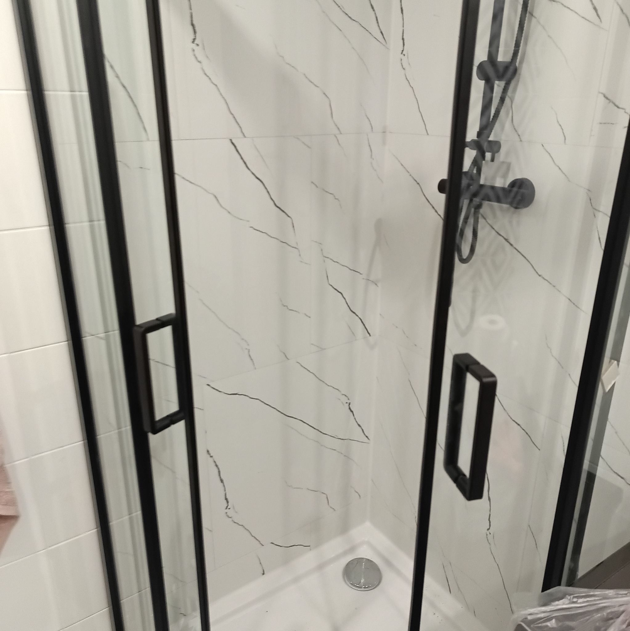 Nowoczesna kabina prysznicowa z czarnymi profilami i armaturą, wykończona białymi płytkami imitującymi marmur. Minimalistyczny design, widoczny odpływ liniowy.