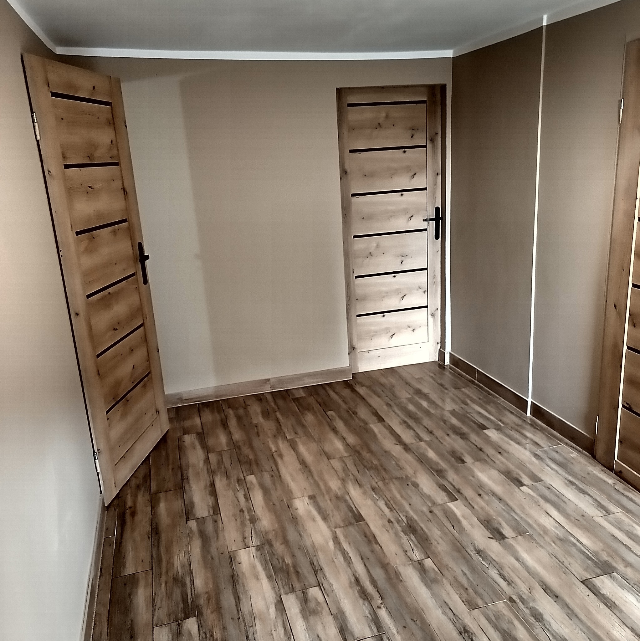 Wykończone wnętrze pokoju z drewnopodobną podłogą, beżowymi ścianami i dwojgiem drzwi z drewnianymi okładzinami i czarnymi, poziomymi wstawkami. Listwy przypodłogowe w kolorze podłogi.