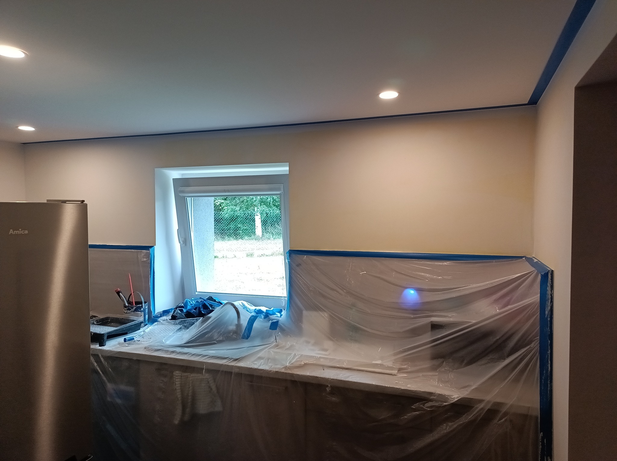 Kuchnia w trakcie malowania: ściany zabezpieczone folią i taśmą malarską, widoczne narzędzia malarskie, sufit z oświetleniem punktowym, przygotowania do malowania.