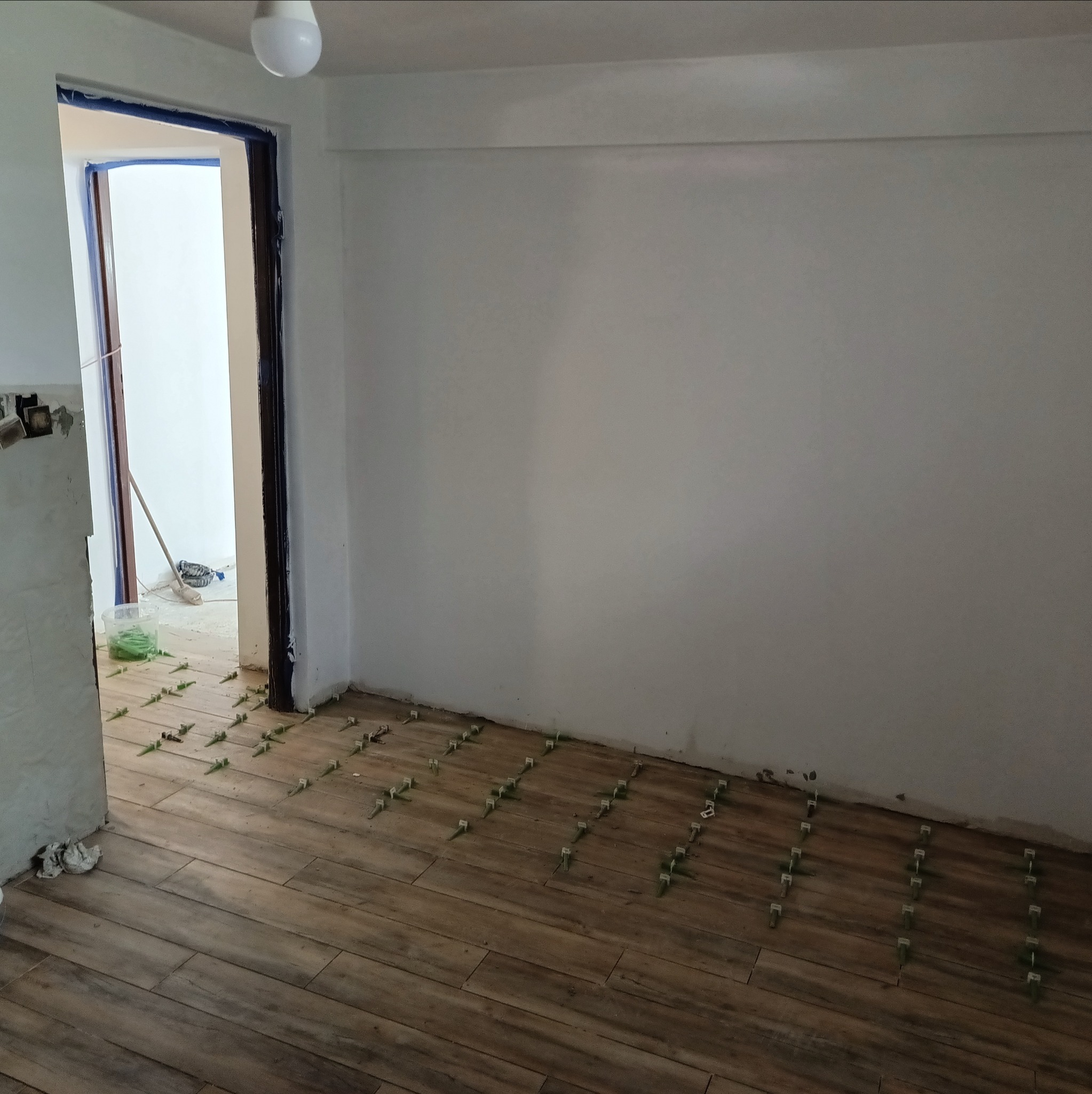 Układanie drewnopodobnych płytek podłogowych z użyciem zielonych krzyżyków dystansowych w jasnym pomieszczeniu. Widoczny fragment ściany i otwarte drzwi.