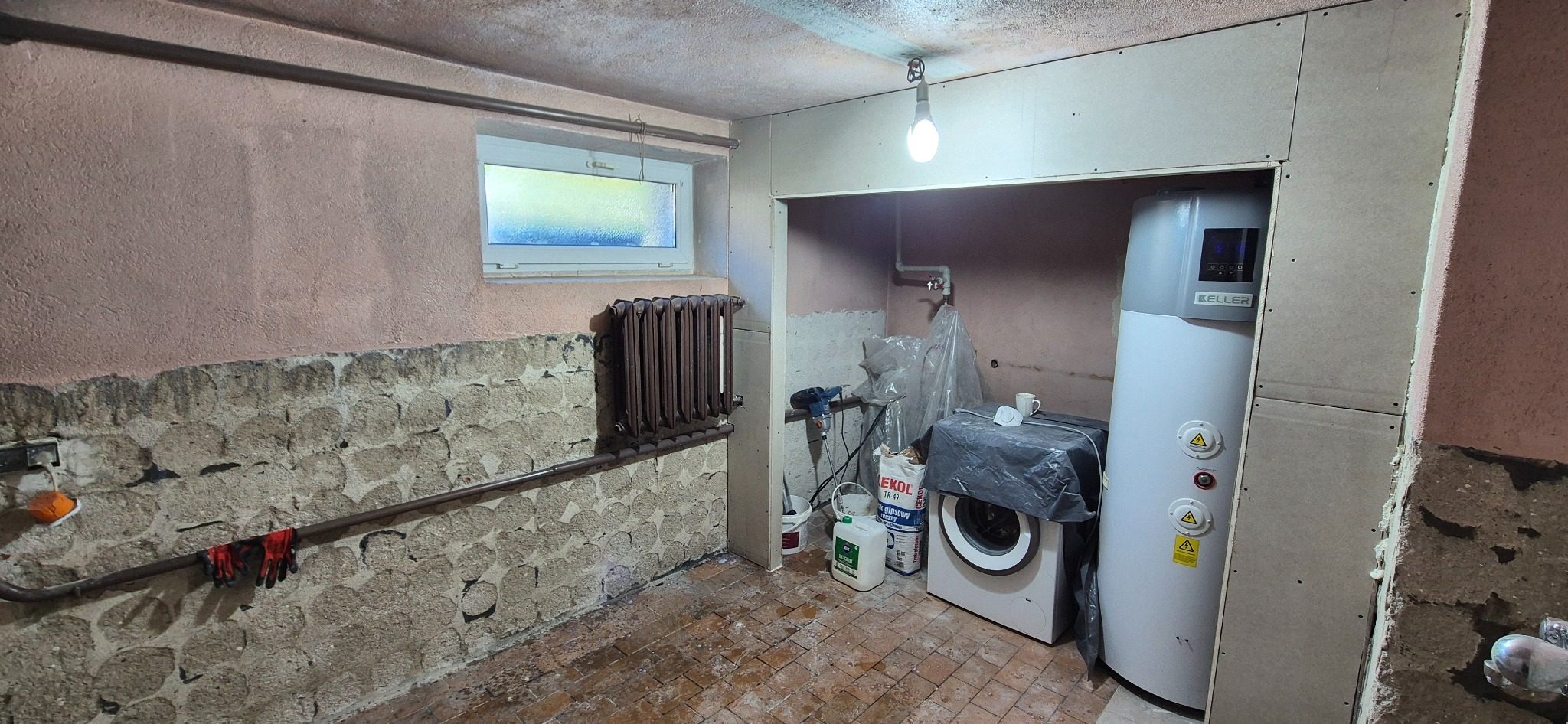 Piwnica w trakcie remontu: ściany z surowego kamienia i częściowo pokryte płytami karton-gips, grzejnik, bojler, pralka, narzędzia i materiały budowlane. Widoczne ślady prac.
