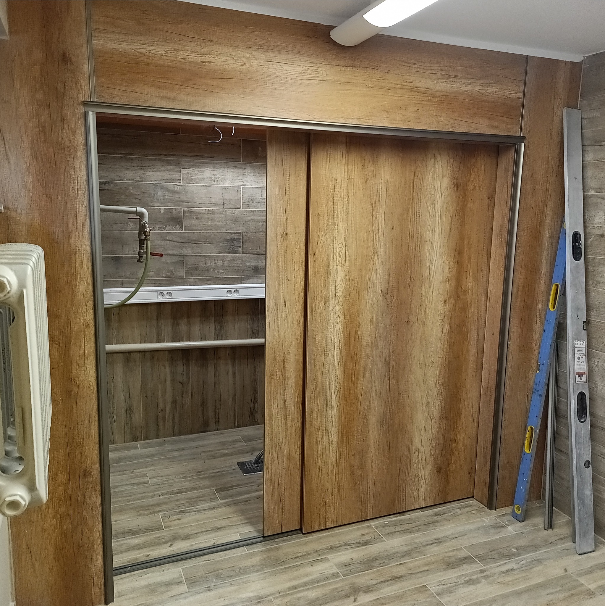 Wykończona łazienka z przesuwnymi drzwiami o fakturze drewna, płytkami imitującymi drewno na ścianach i podłodze. Widoczny grzejnik i poziomice.