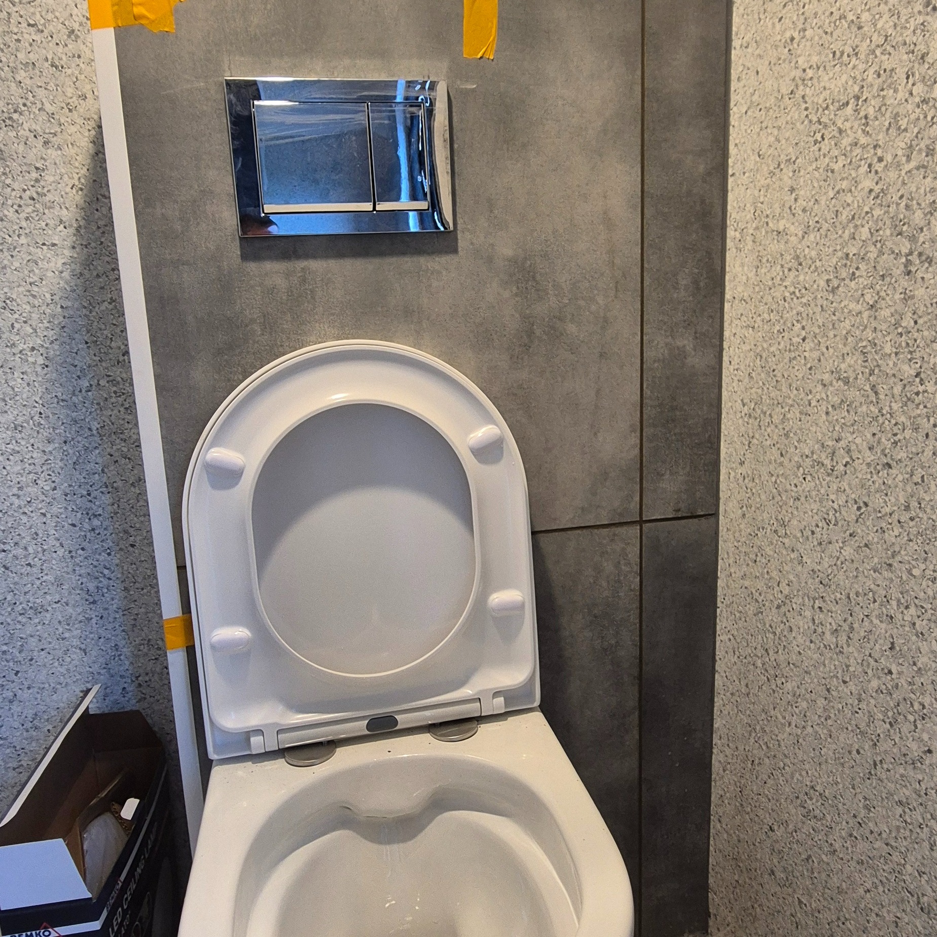 Zamontowana toaleta wisząca z chromowanym przyciskiem spłukującym na szarej ścianie z płytek. Widoczne taśmy malarskie i karton po oświetleniu LED.