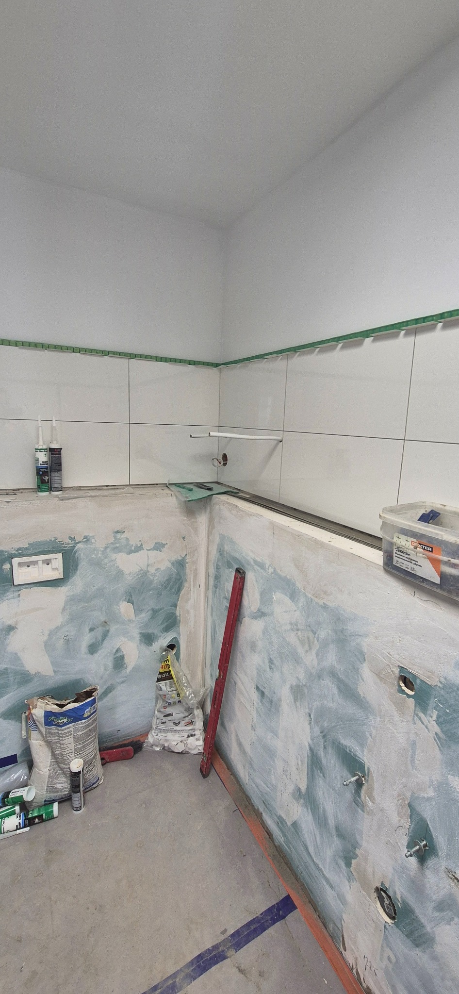 Remont łazienki w trakcie: częściowo położone białe płytki z zieloną taśmą, ściany przygotowane do dalszych prac, widoczne narzędzia i materiały budowlane.