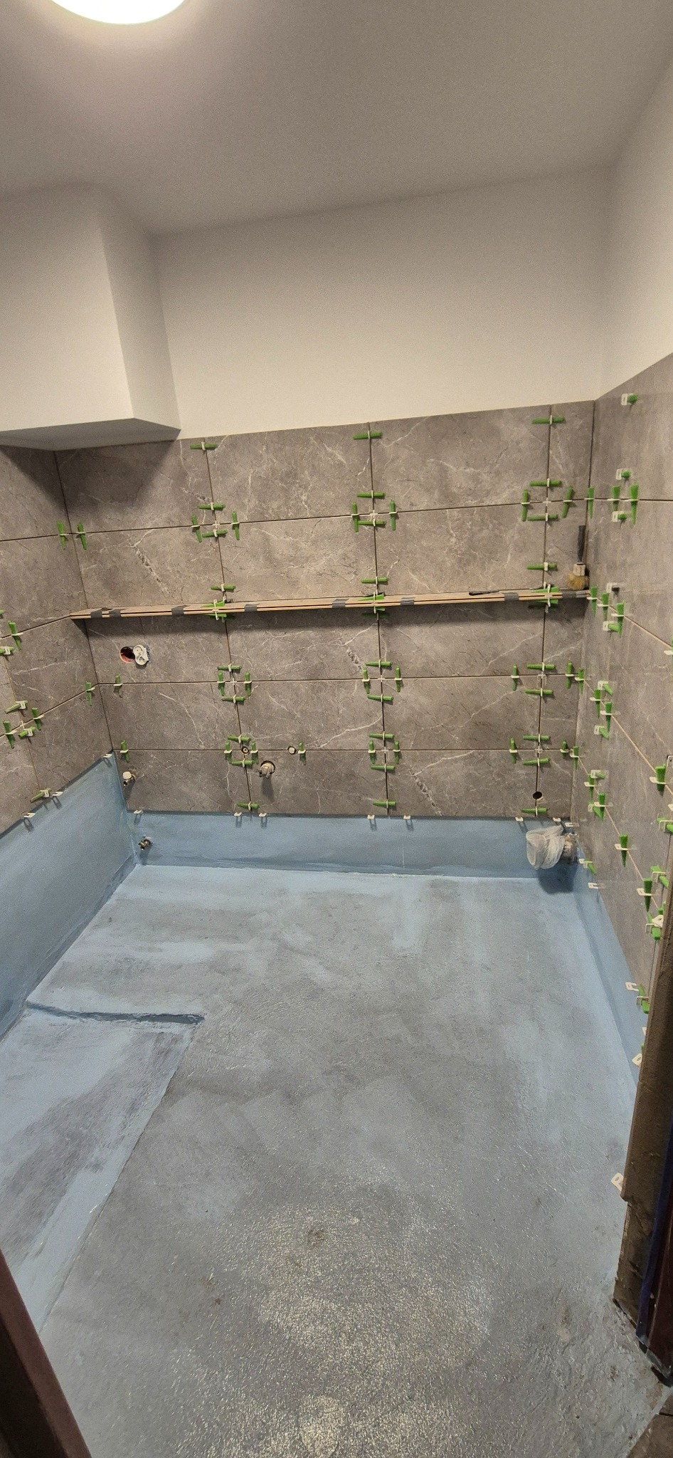 Łazienka w trakcie prac: szare płytki z zielonymi krzyżykami dystansowymi na ścianach, podłoga pokryta niebieską hydroizolacją. Widoczny etap układania płytek.