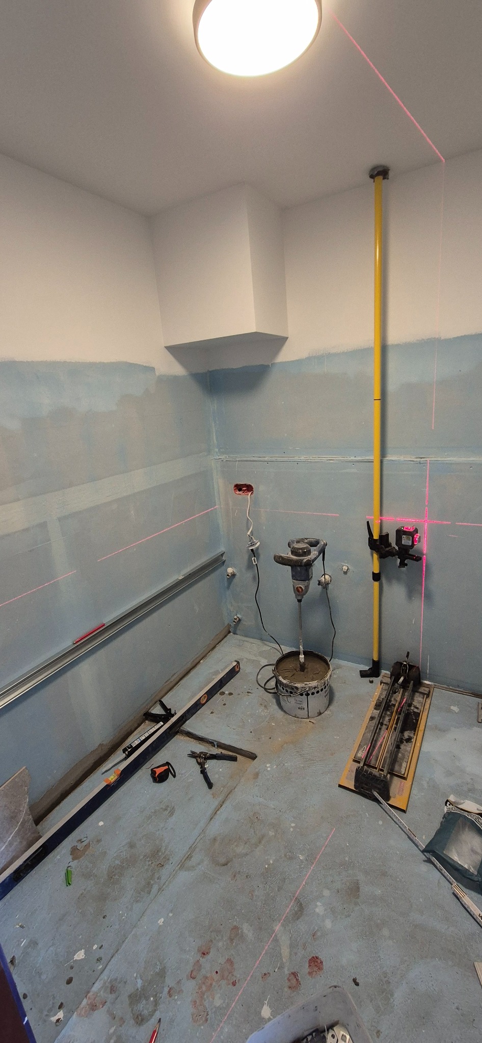 Przygotowanie do układania płytek w łazience: poziomica laserowa, mieszadło do zaprawy, narzędzia glazurnicze. Ściany częściowo pokryte gruntem, widoczne ślady po wyznaczaniu poziomu.