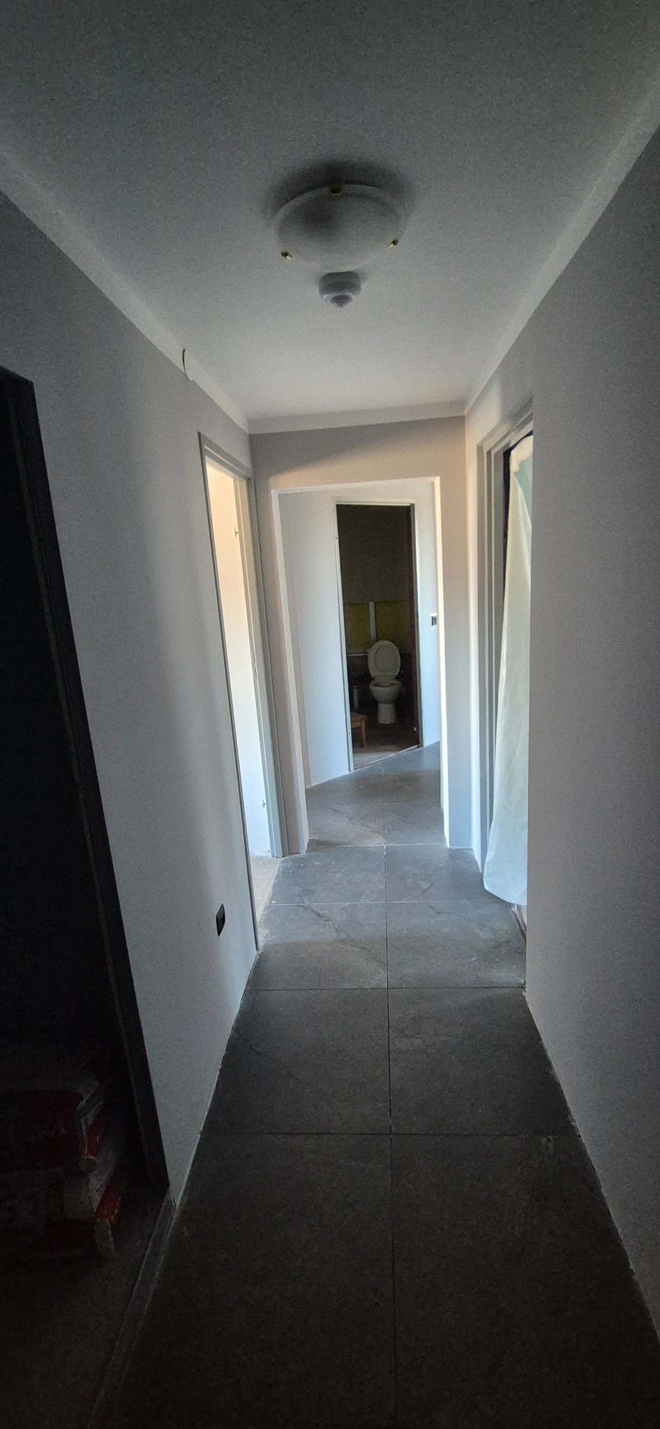 Wykończony korytarz z szarą podłogą z płytek i świeżo pomalowanymi ścianami na szaro. W tle widoczna łazienka z toaletą. Nowoczesne oświetlenie sufitowe.