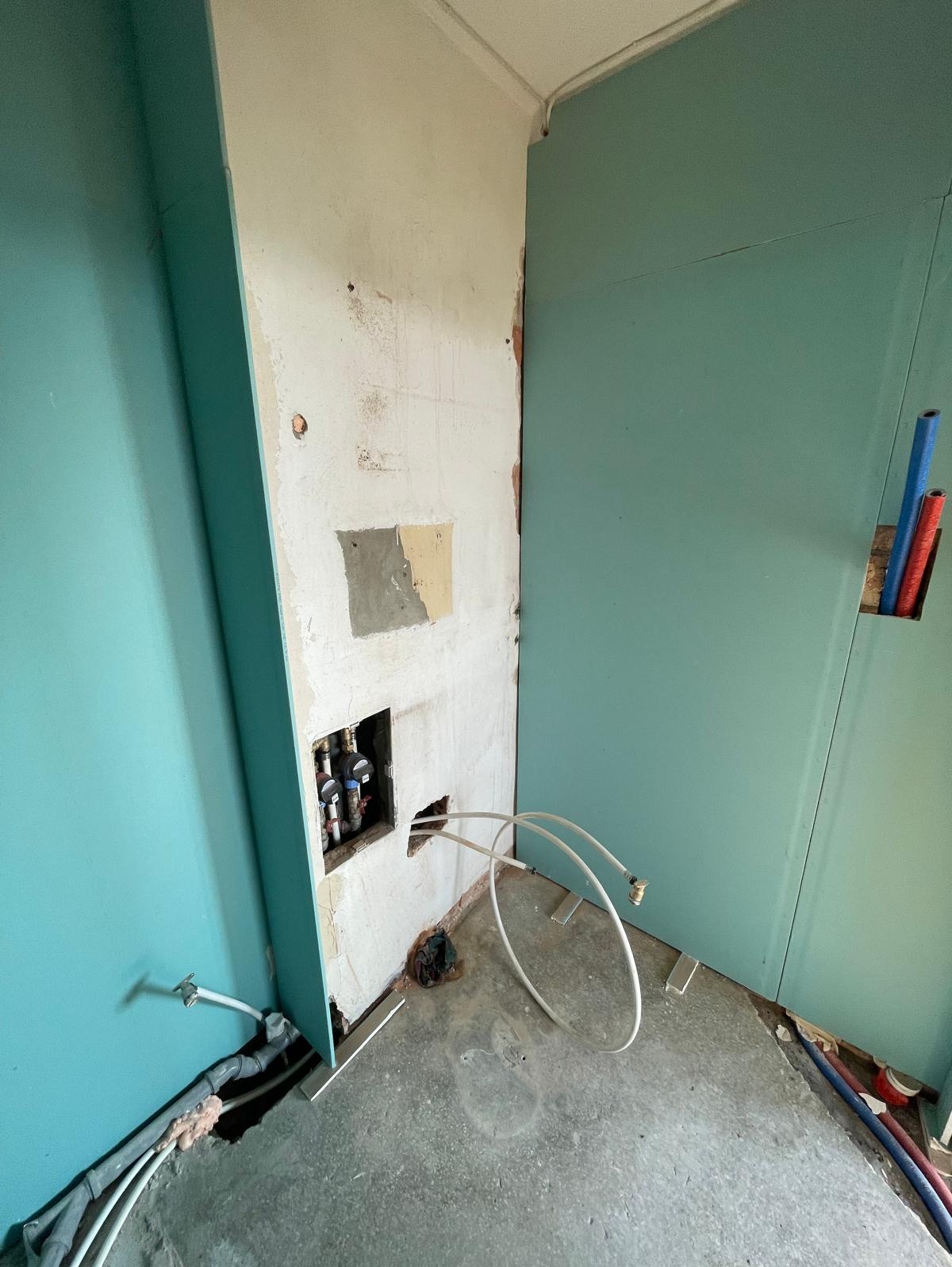 Narożnik łazienki w trakcie prac: płyty GK w kolorze turkusowym, fragment ściany z instalacją wodną i wystającymi rurkami. Surowa podłoga, widoczne przewody.
