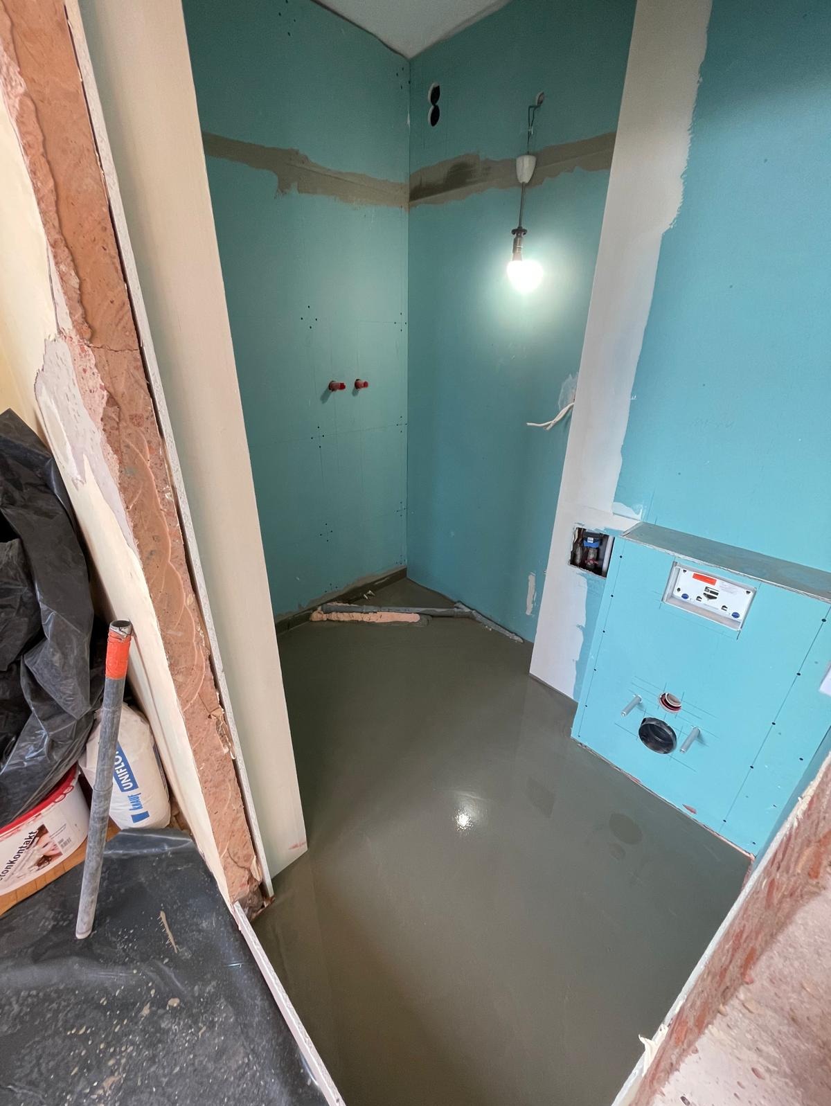 Wnętrze łazienki w trakcie prac: wylewka podłogowa, zabudowa stelaża WC z płyty GK, ściany z płyt GK przygotowane do dalszych prac wykończeniowych. Widoczne materiały budowlane.