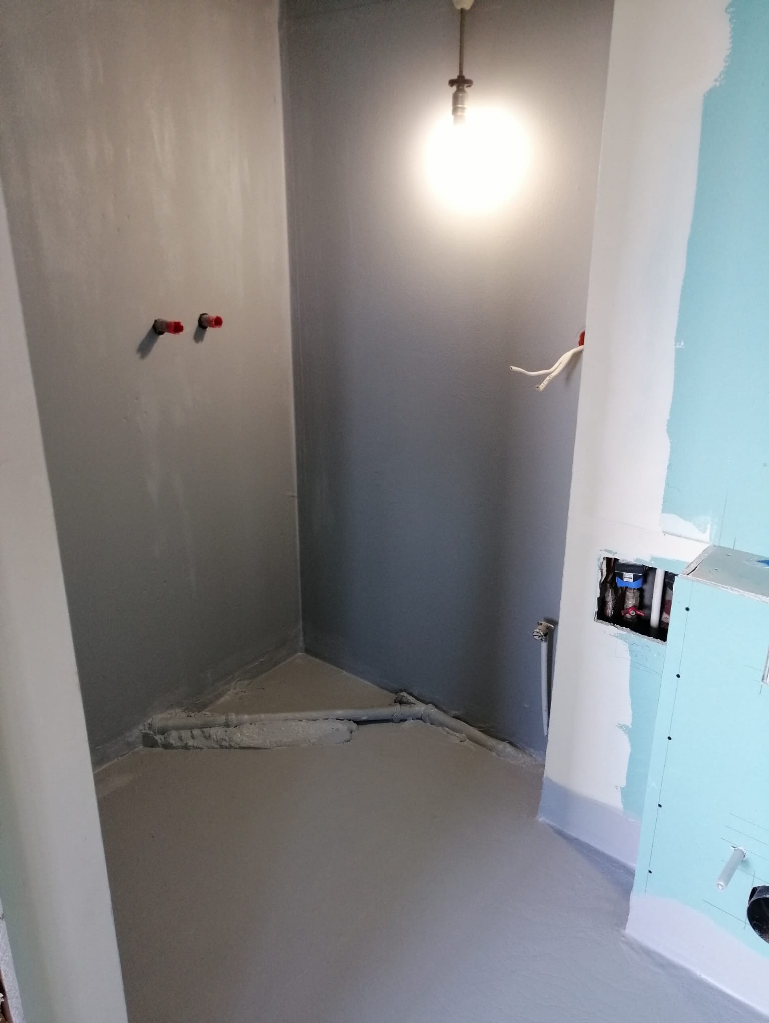 Szara łazienka w trakcie prac: wylewka na podłodze, ściany przygotowane pod płytki, instalacja wodna. Widoczne rury i otwór w ścianie.