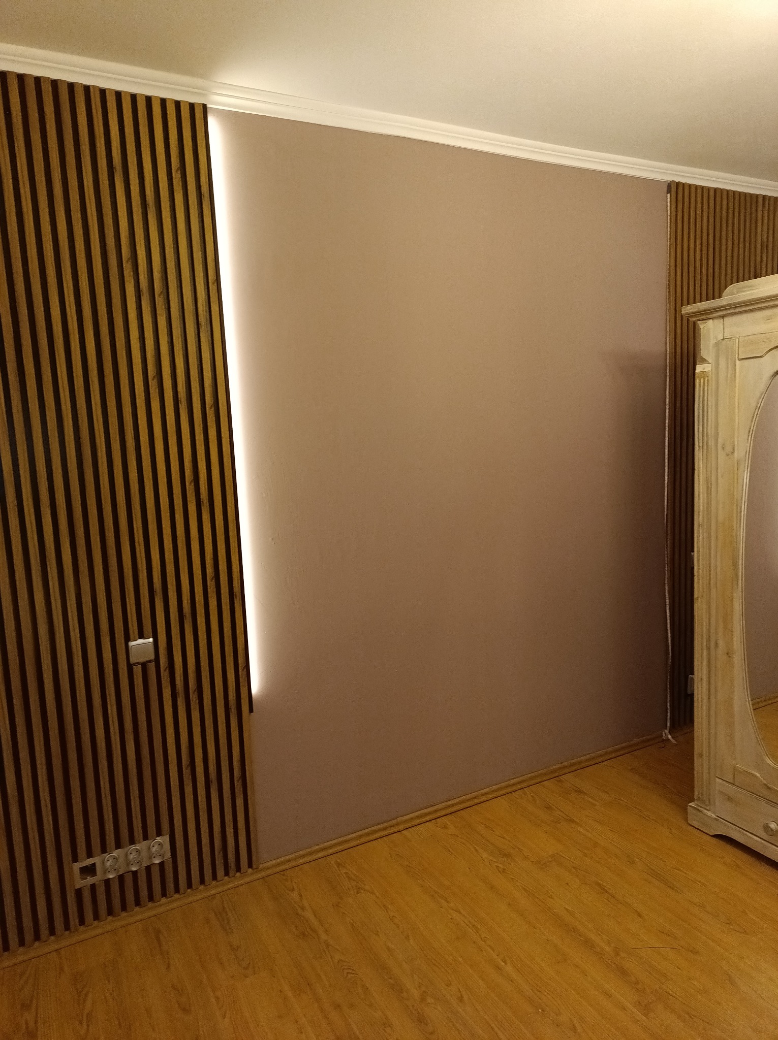 Beżowa ściana z drewnianymi lamelami i ukrytym oświetleniem LED. Widoczny fragment podłogi i szafy. Nowoczesny design wnętrza.