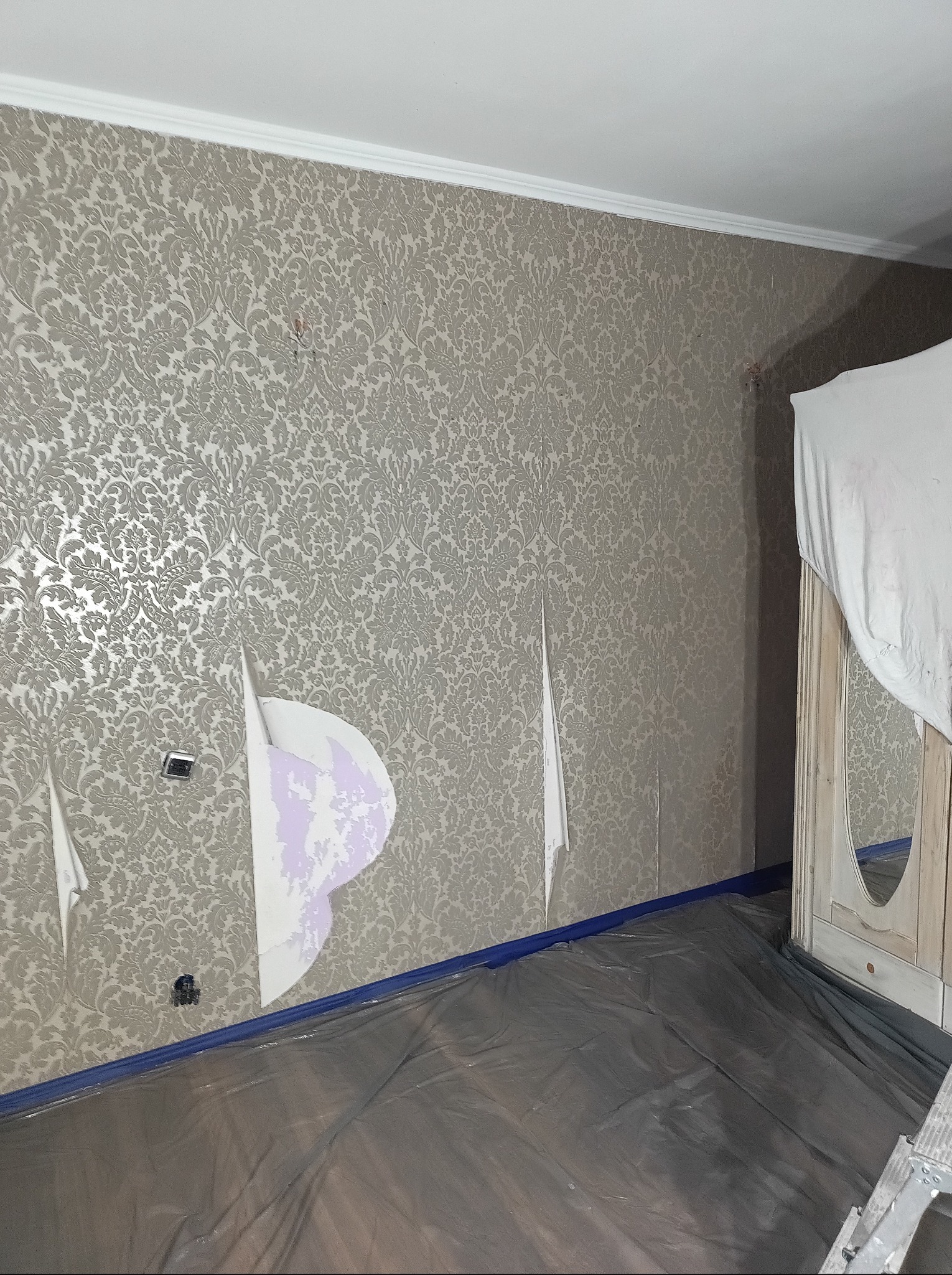 Pokój w trakcie usuwania starej tapety z wzorem. Fragmenty tapety odklejone, widoczne ślady kleju. Podłoga zabezpieczona folią malarską. Przy ścianie stoi szafa.