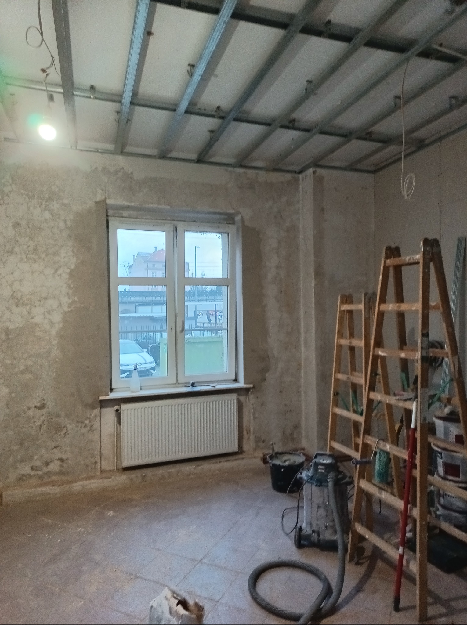 Wnętrze w trakcie prac w Kłodzku: surowe ściany, metalowa konstrukcja sufitu podwieszanego, drabiny, odkurzacz przemysłowy. Widok na okno i budynek na zewnątrz.