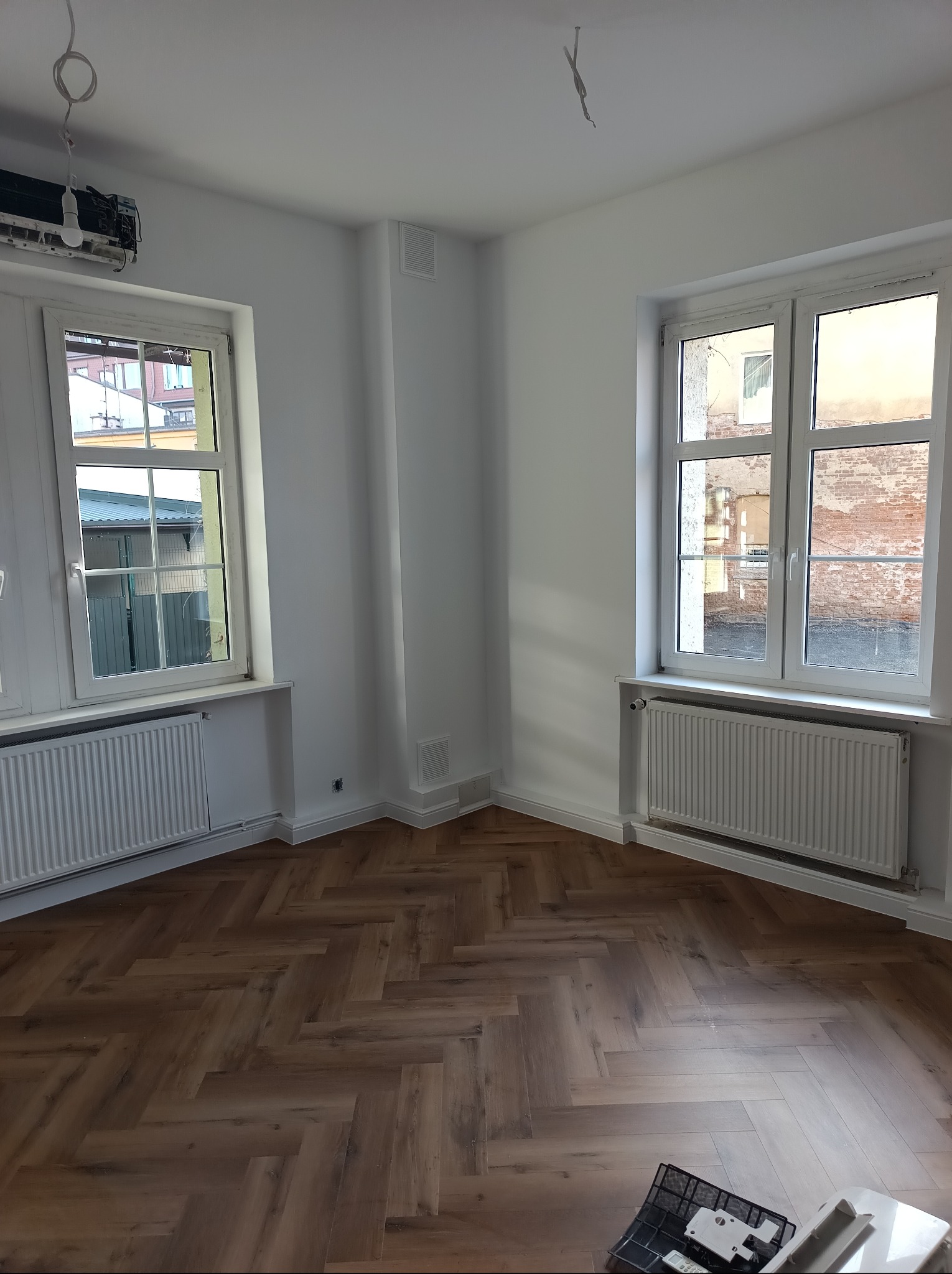 Jasne wnętrze po remoncie: parkiet w jodełkę, białe ściany, okna z grzejnikami. Widoczne niedokończone detale instalacji elektrycznej i akcesoria montażowe.