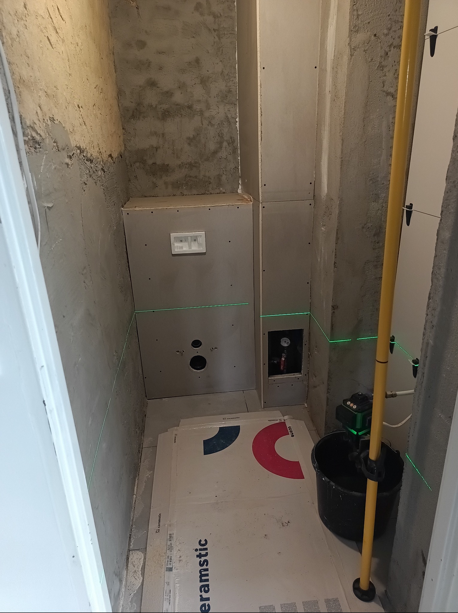 Przygotowanie do układania płytek w małej łazience: surowe ściany, płyty gipsowe, laser krzyżowy i paczki płytek ceramicznych na podłodze. Widok z góry.